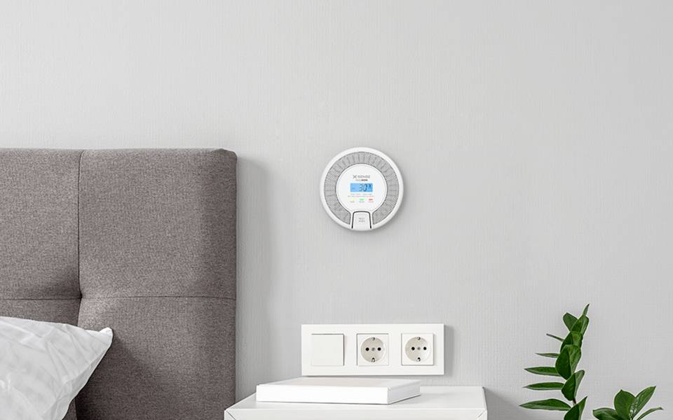 Ein intelligentes Thermostat, das Temperatur und Einstellungen anzeigt, ist an einer grauen Wand über einem Kopfteil und neben einer weißen Steckdose montiert.