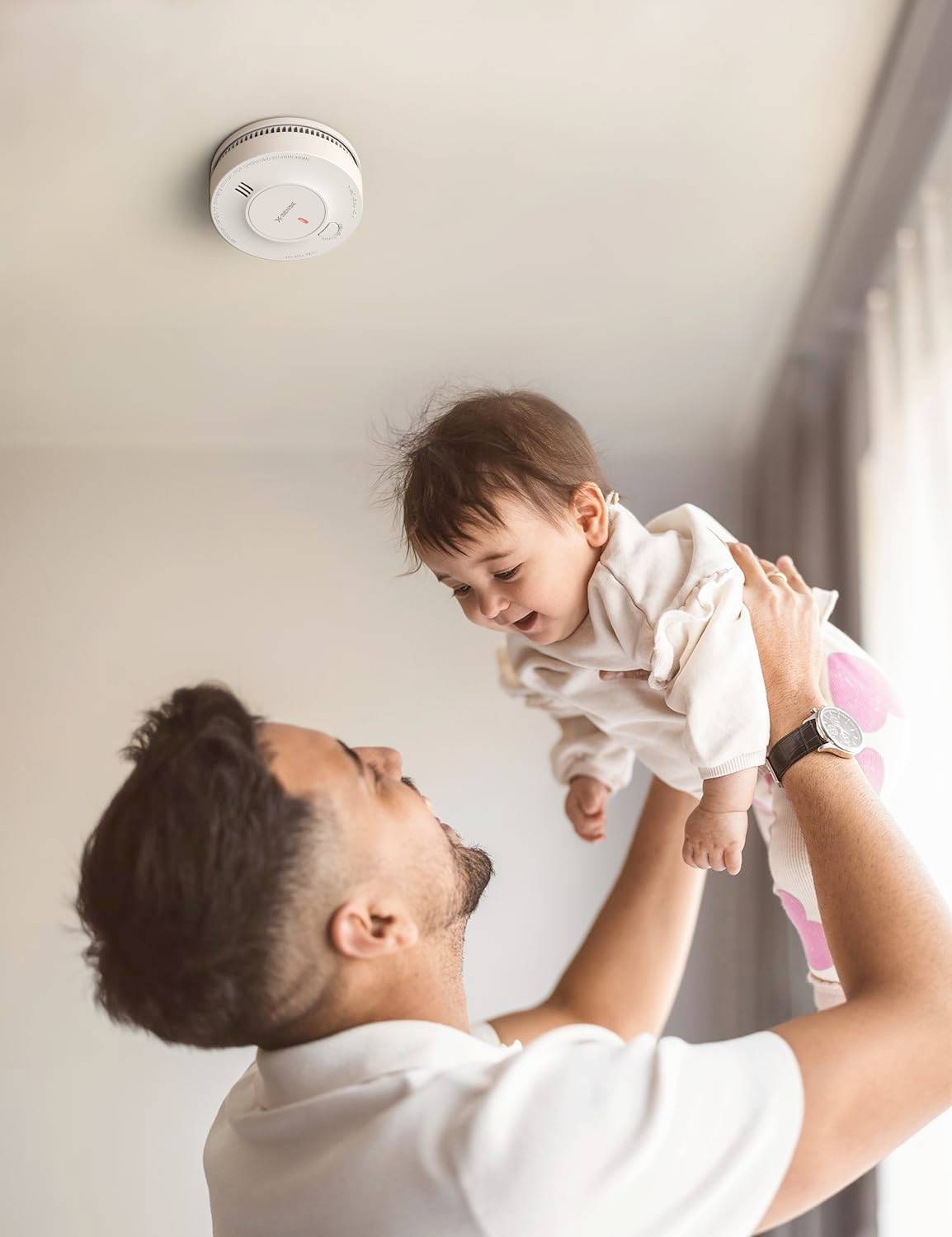 Eine Person hält ein lächelndes Baby zur Decke empor, wo ein Rauchmelder installiert ist und damit die Sicherheit im Haus betont.