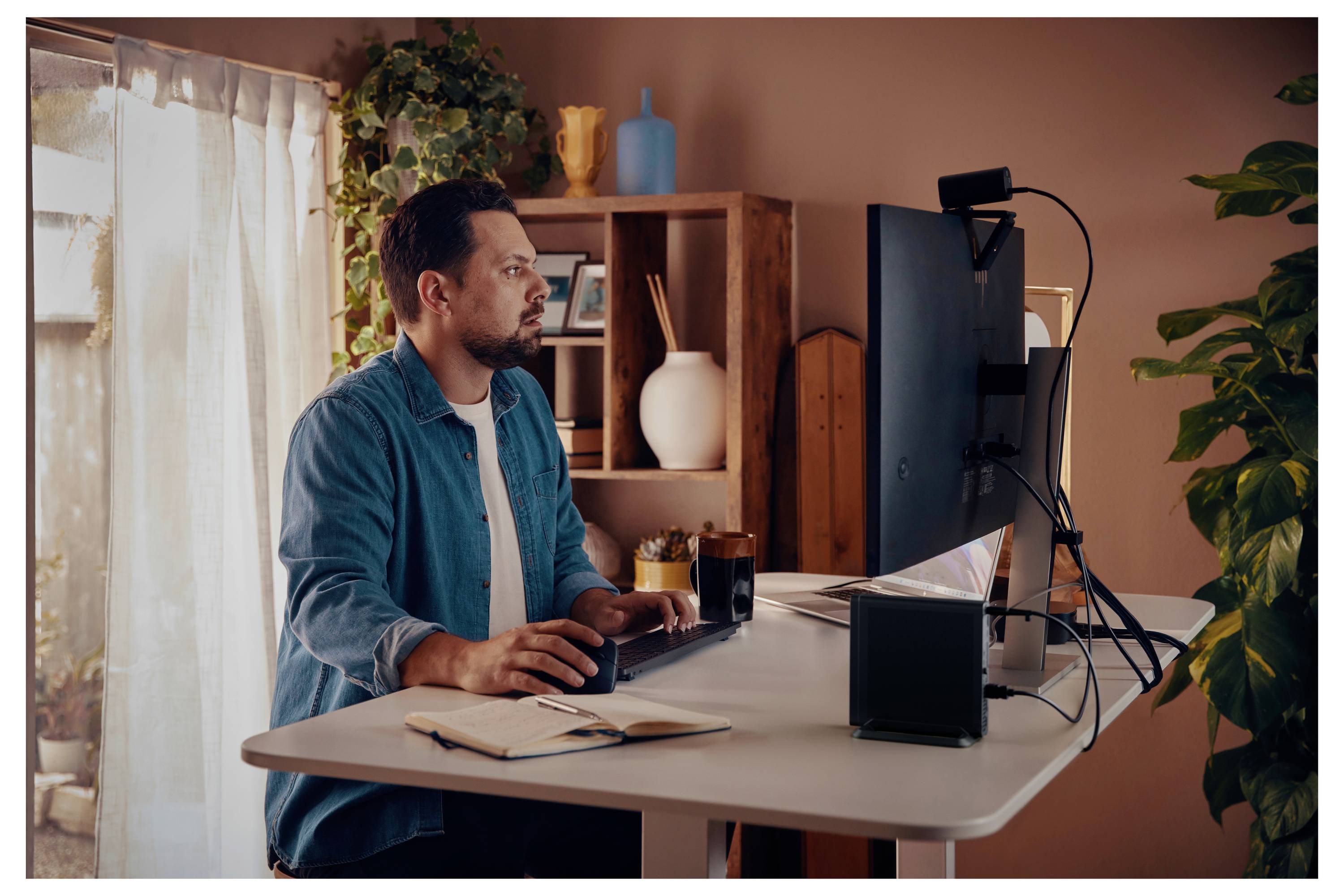 Ein Mann, der an einem Stehschreibtisch mit zwei Monitoren arbeitet, umgeben von Pflanzen und Büchern. Sonnenlicht filtert durch einen Vorhang und schafft eine warme Atmosphäre.
