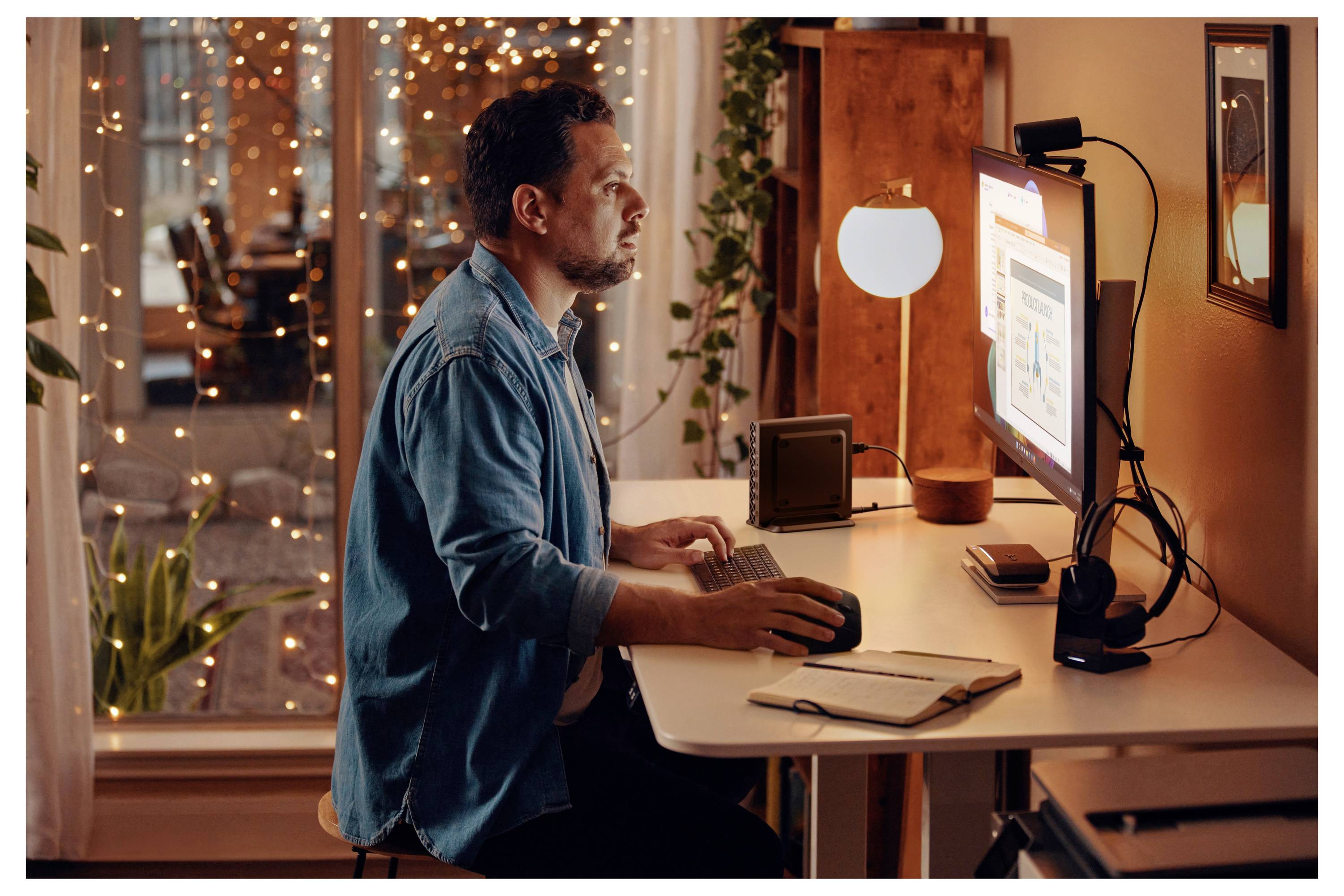 Ein Mann sitzt an einem Schreibtisch und arbeitet an einem Computer, konzentriert auf den Bildschirm, der eine Präsentation zeigt. Warmes Licht und Lichterketten schaffen eine gemütliche Atmosphäre.