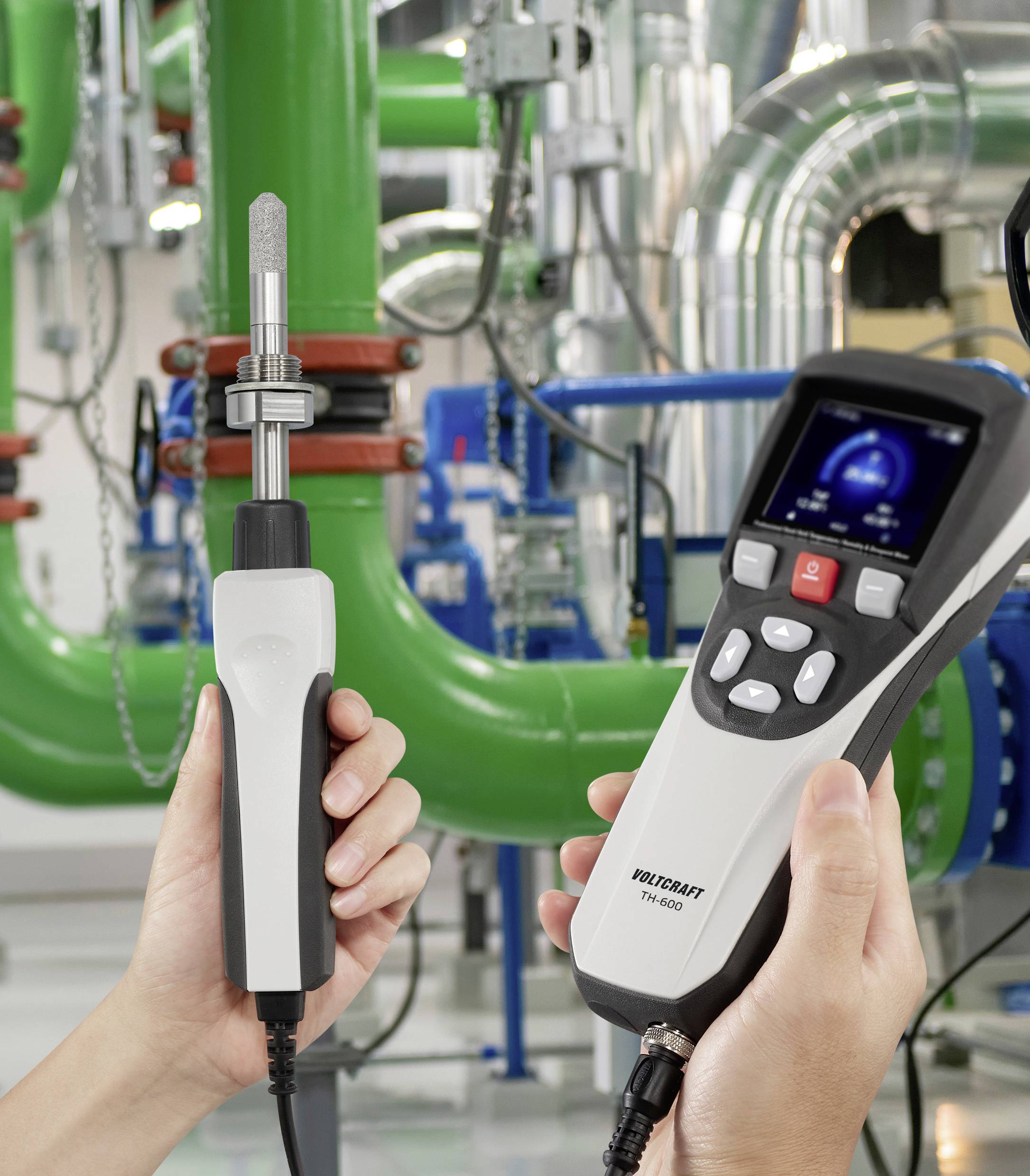 Eine Person hält ein digitales Thermometer und eine Sonde in einer Industrieanlage mit grünen Rohren und Maschinen im Hintergrund.