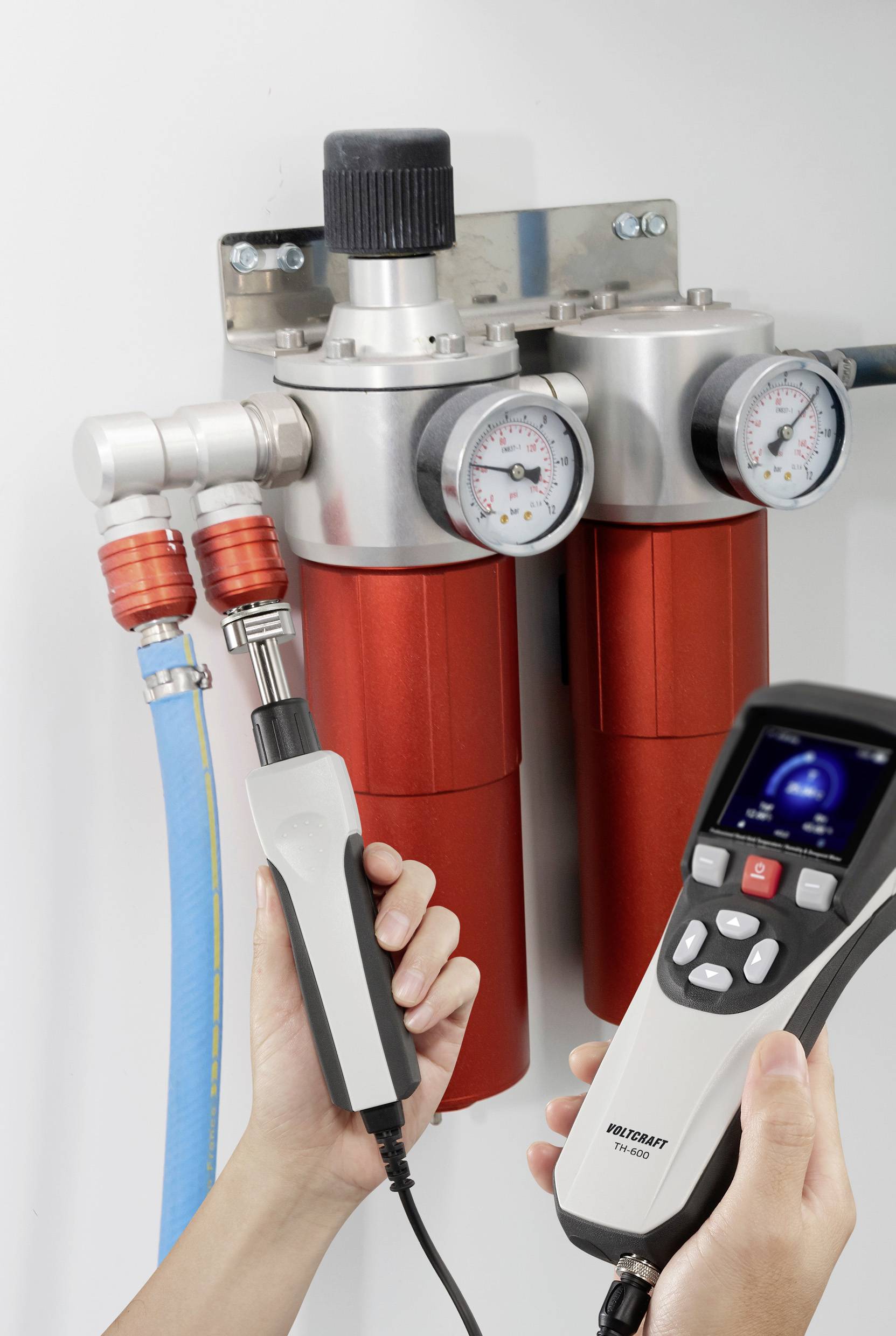 Eine Person nutzt ein tragbares Gerät, um den Druck von zwei roten zylindrischen Behältern mit Druckmessern zu messen, die durch Gummischläuche verbunden sind.