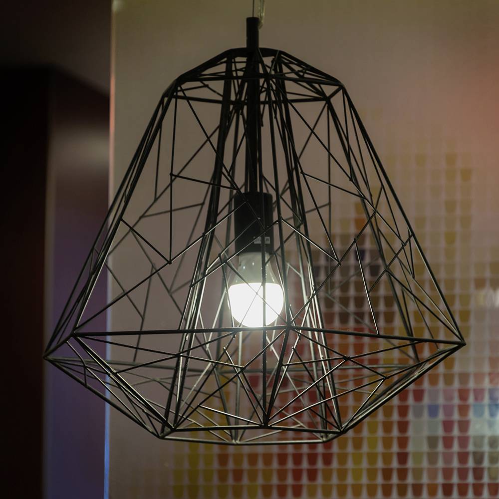 Eine geometrische Metall-Pendelleuchte, die von der Decke hängt, mit einer leuchtenden Glühbirne im Inneren, die komplexe Schatten auf strukturierter Tapete wirft.