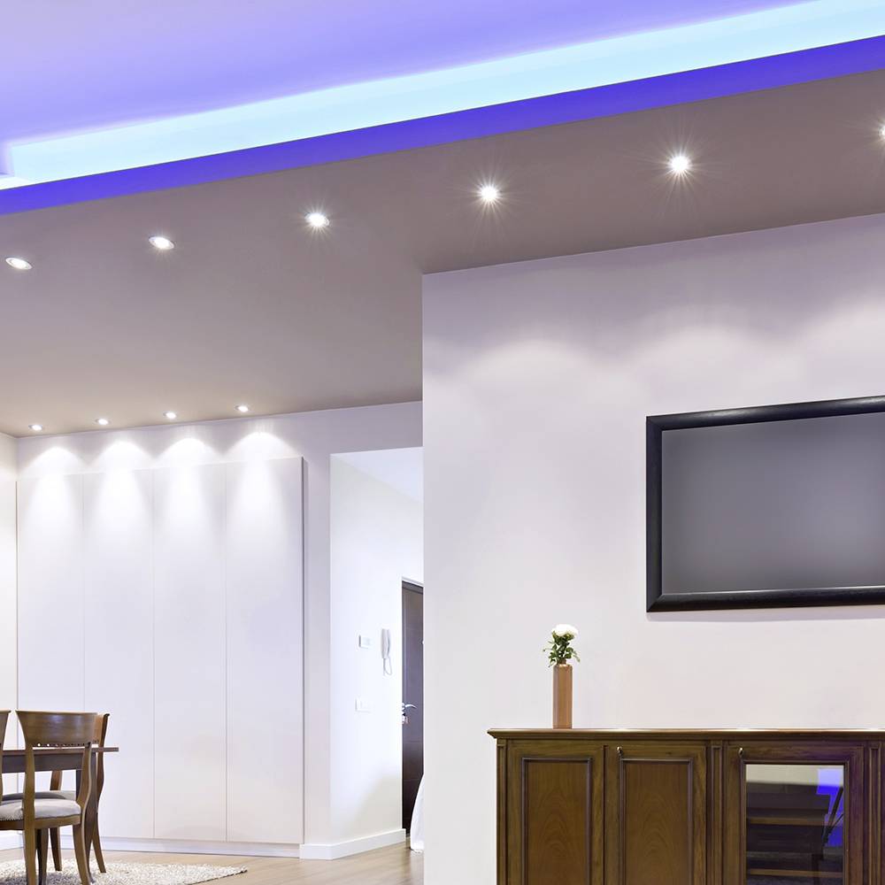Ein moderner Wohnraum mit Einbauleuchten, einem wandmontierten Fernseher und Holzmöbeln darunter. Hell erleuchtete Decke mit blauen Akzenten.