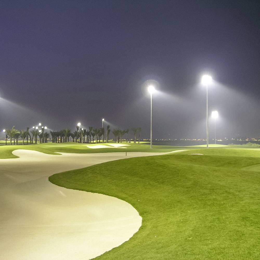 Ein Golfplatz bei Nacht, beleuchtet von hellen Lichtern, mit sattgrünen Fairways und Sandbunkern, umgeben von Palmen.
