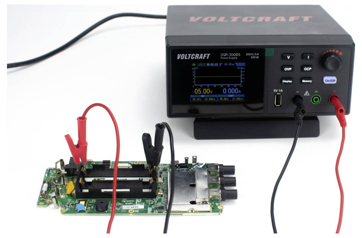Eine digitale Stromversorgungseinheit, die über Kabel mit einer Leiterplatte verbunden ist. Das Display zeigt Spannungsmessungen, die auf eine aktive Prüfung oder Kalibrierung hindeuten.