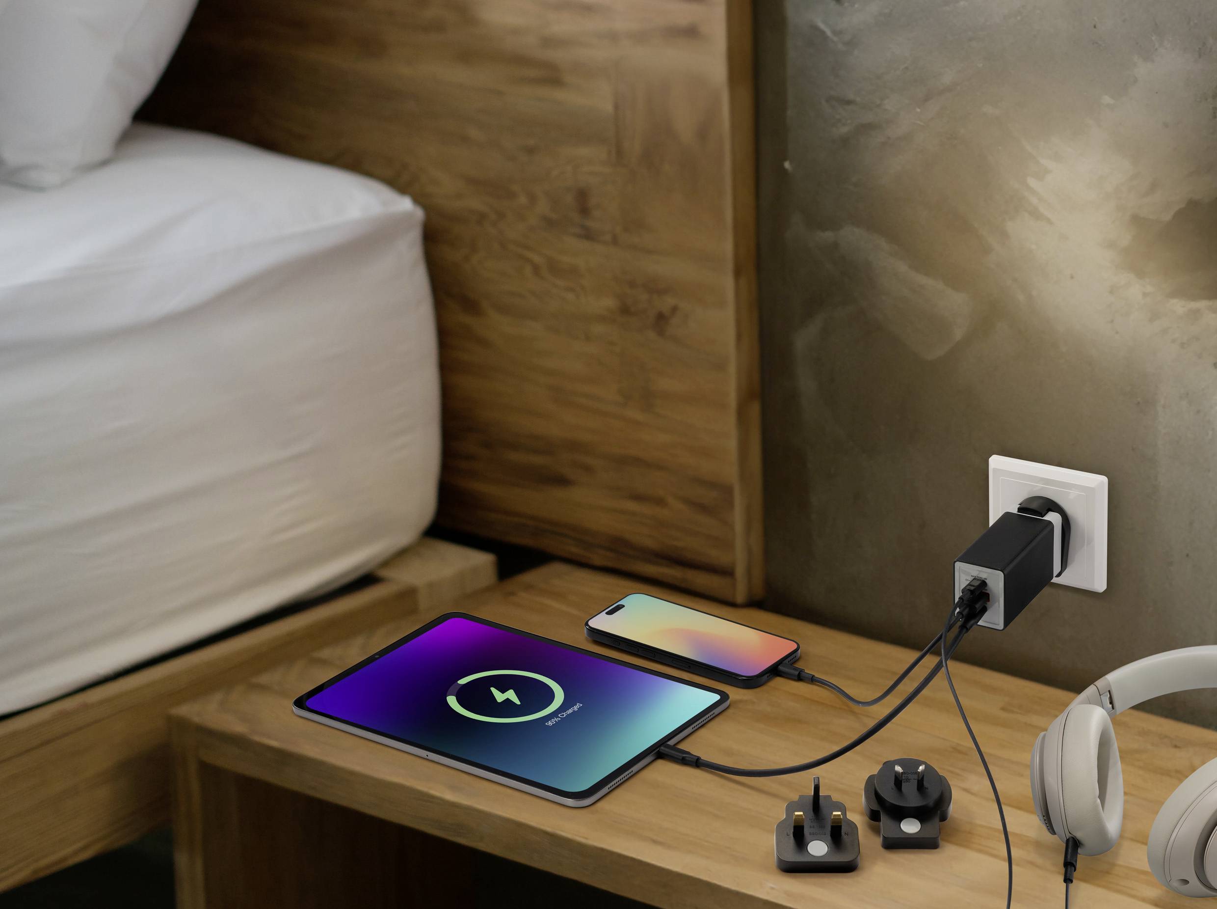 Tablet und Smartphone laden auf einem Holznachttisch mit einem Stecker mit internationalen Adaptern; Kopfhörer liegen in der Nähe.