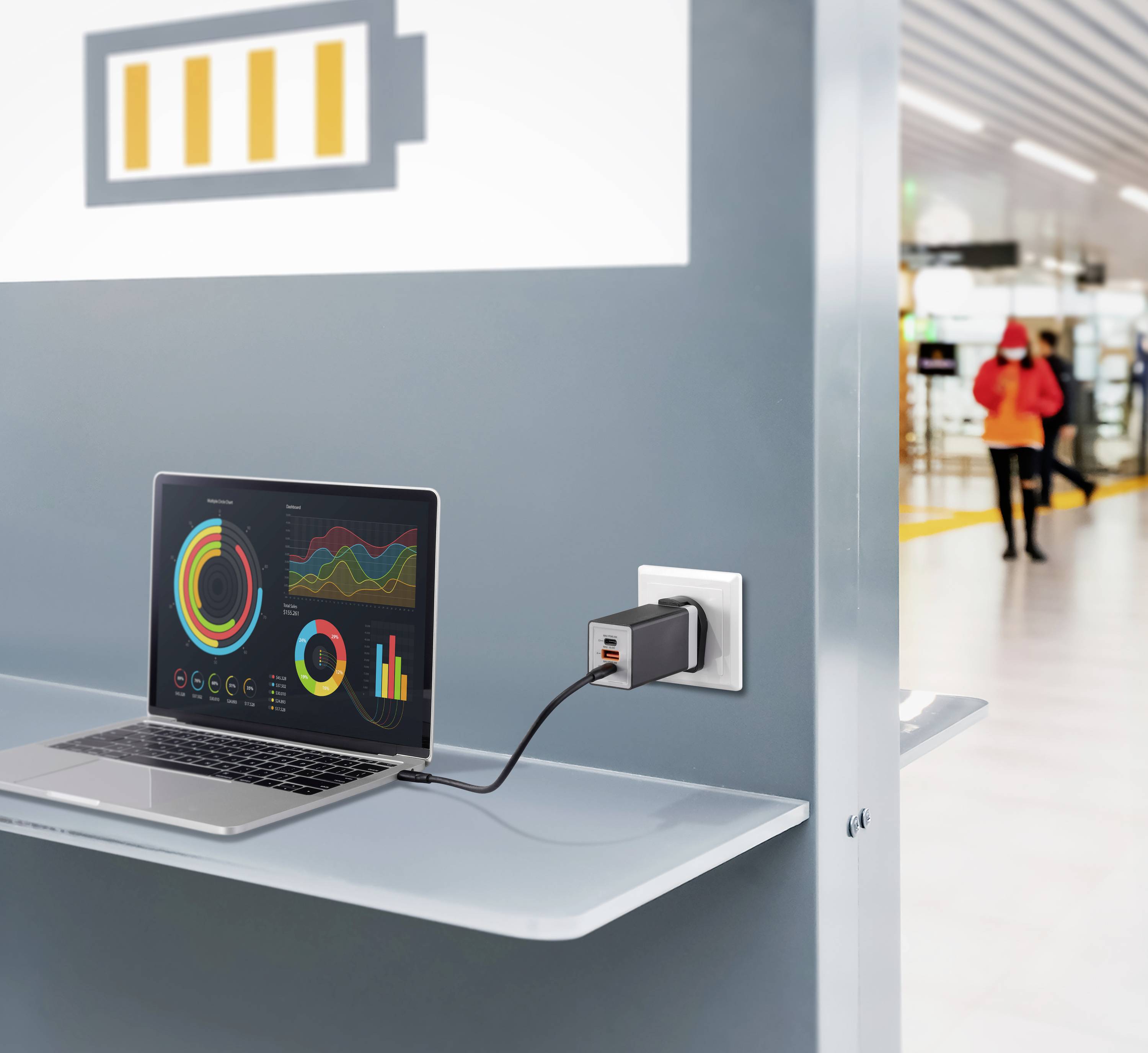 Ein Laptop an einer Ladestation in einer Flughafenlounge, auf dessen Bildschirm farbenfrohe Grafiken und Diagramme zu sehen sind. Eine Steckdose und ein Stecker sind sichtbar.