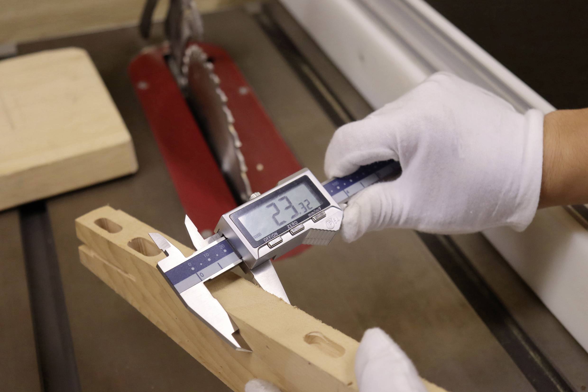 Eine Person mit Handschuhen verwendet eine digitale Schieblehre, um die Breite eines Holzstücks in der Nähe einer Sägeblatt zu messen, wobei die Anzeige '23,72' zeigt.