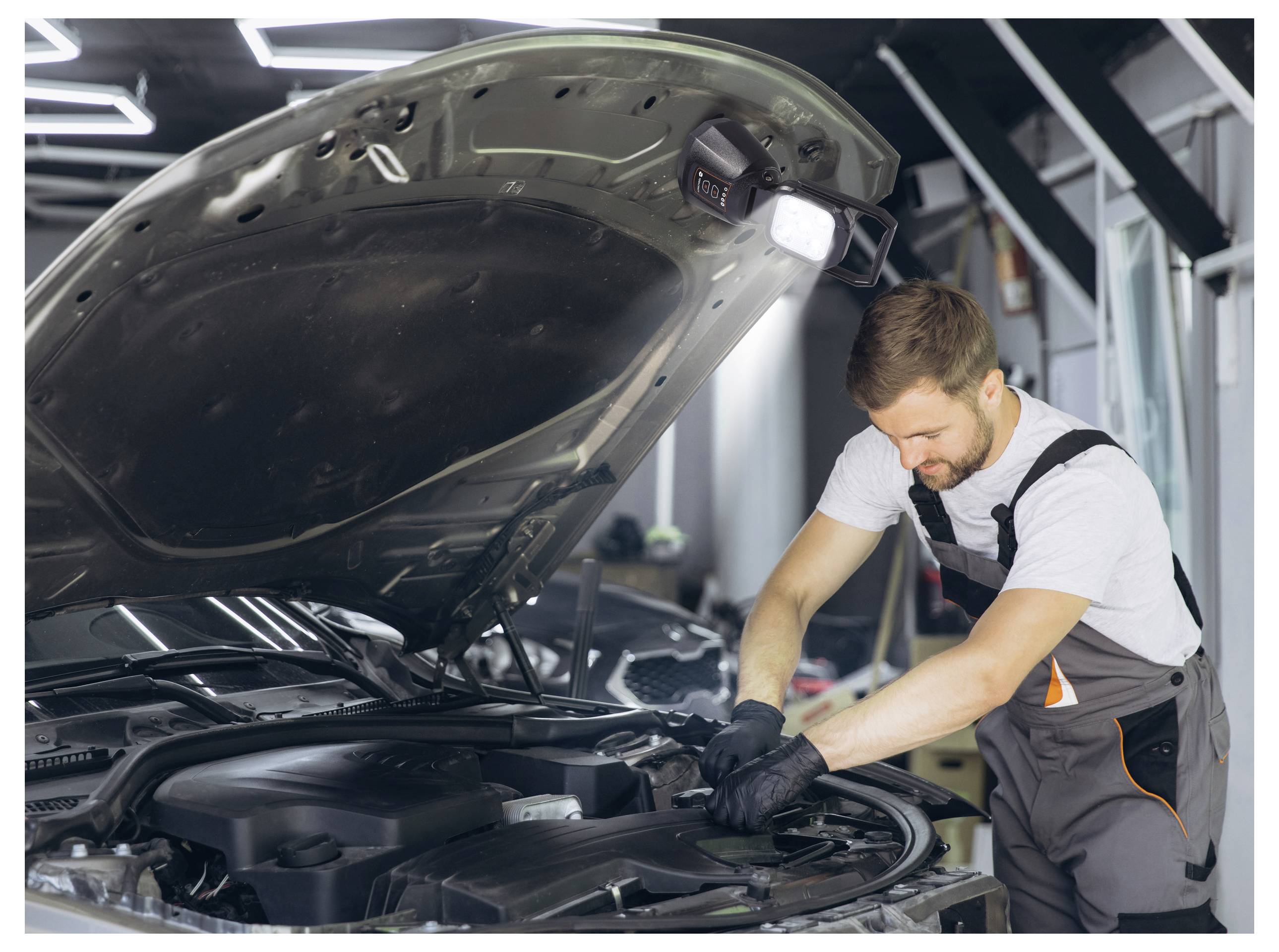 Ein Mechaniker in einer Werkstatt arbeitet an einem Automotor mit offener Motorhaube, trägt Arbeitshandschuhe und Overall und ist konzentriert auf eine Reparaturaufgabe fixiert.