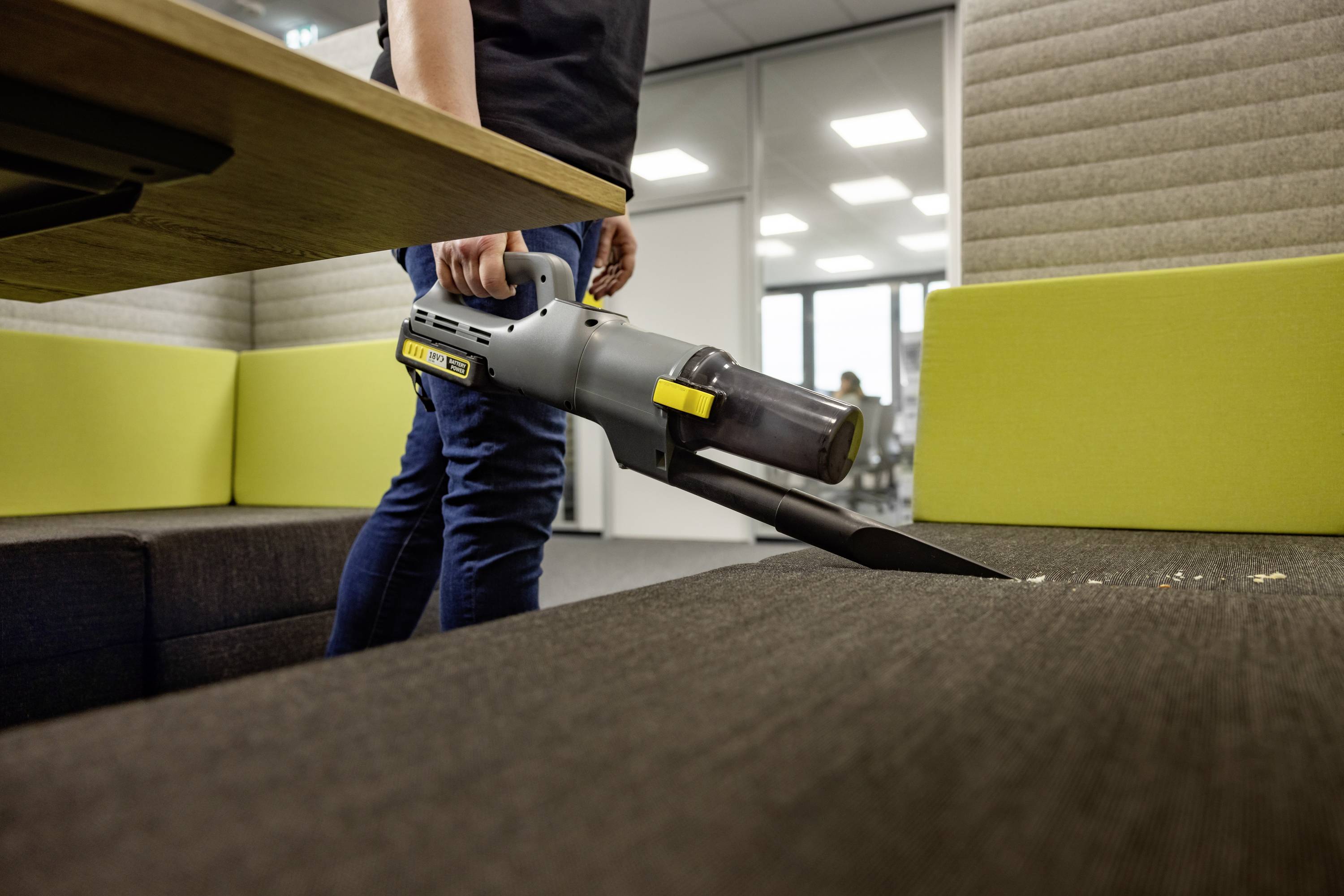 Eine Person benutzt einen tragbaren Staubsauger, um Krümel von einem dunkel gefärbten Sofa in einem modernen Büroumfeld zu reinigen.