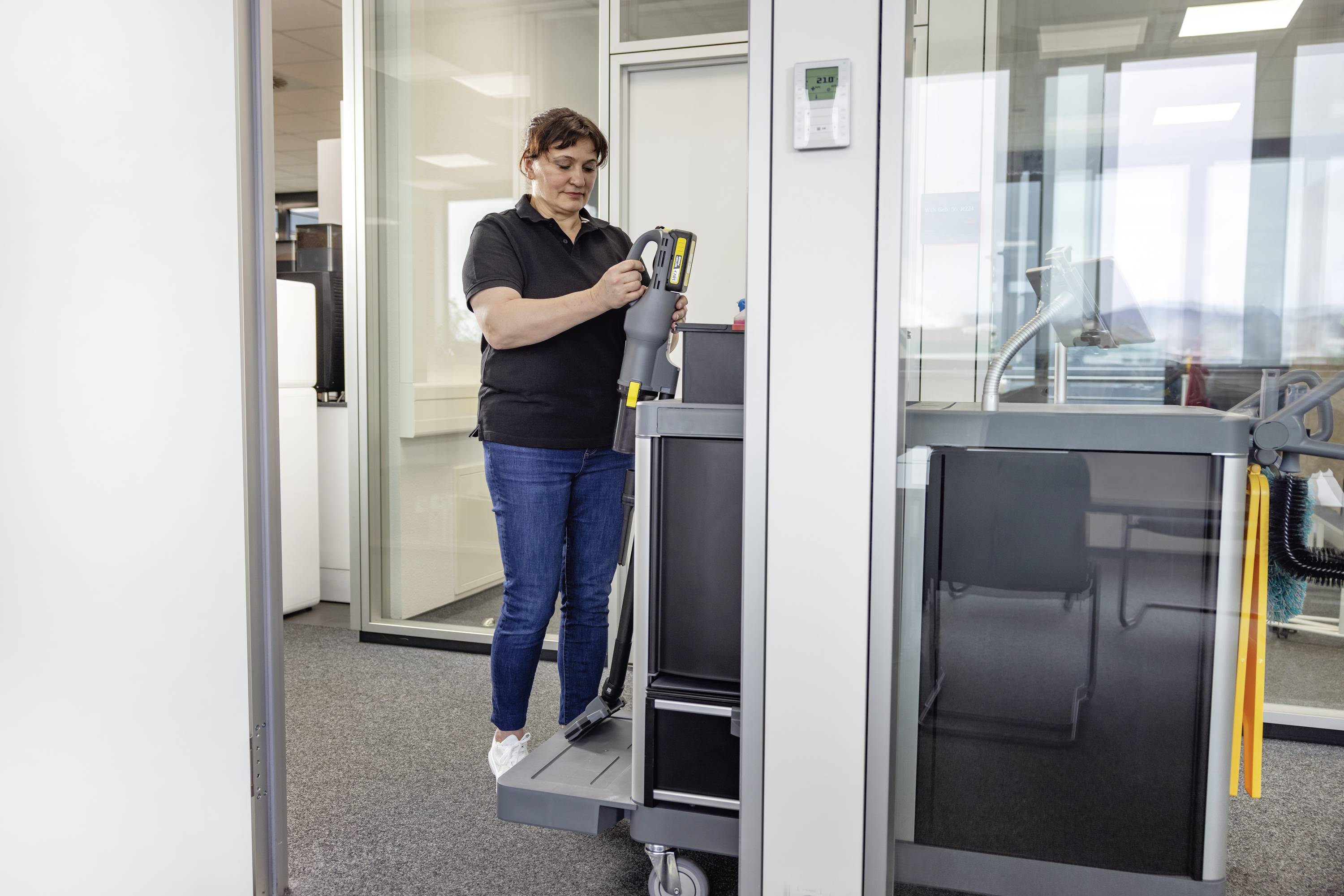 Eine Person in Freizeitkleidung, die neben einem Reinigungswagen in einem Büro steht und ein Reinigungswerkzeug hält, bereit, Aufgaben zu erledigen.