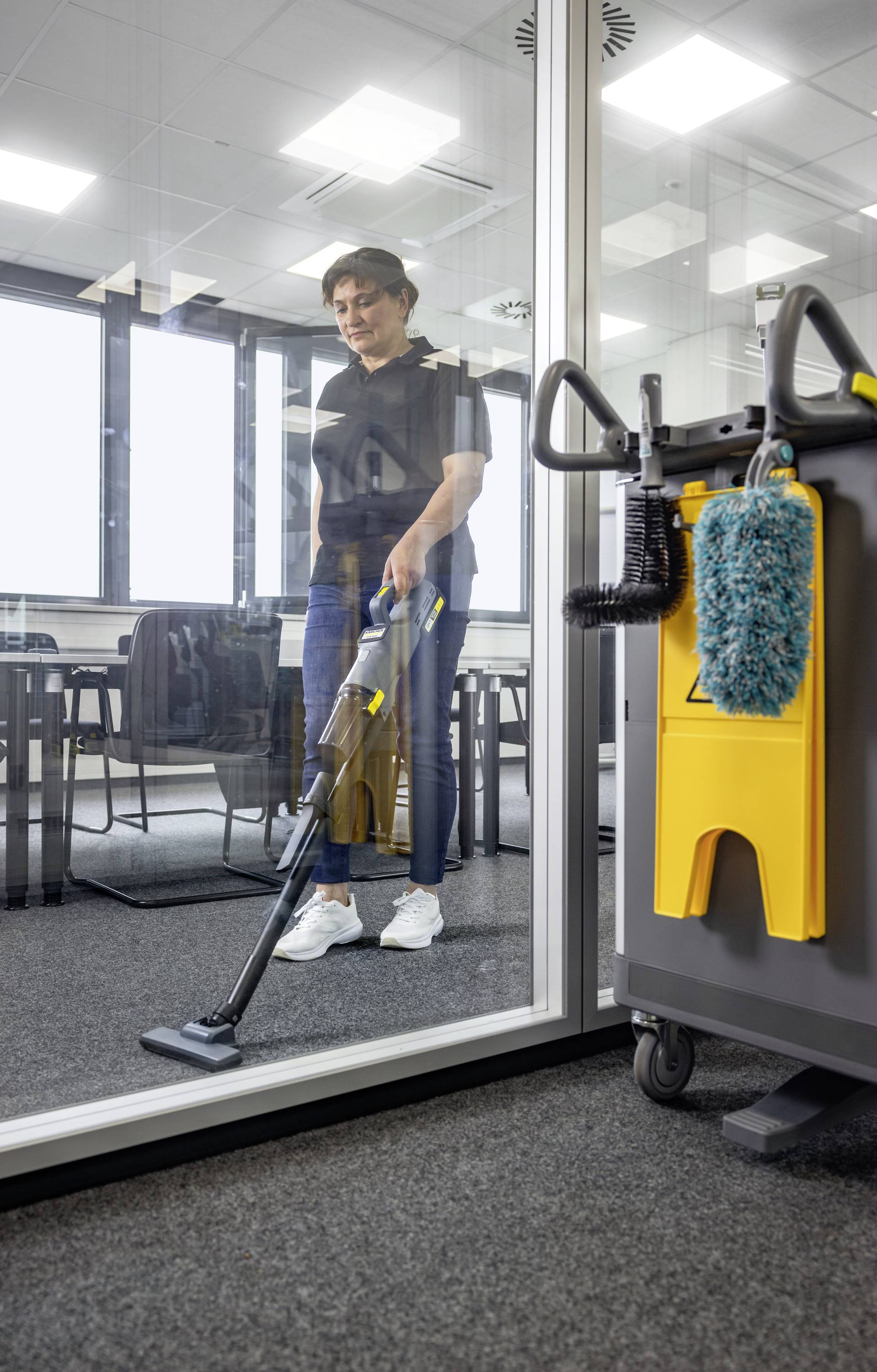 Eine Person, die einen Teppichboden in einem Büroraum mit einem Staubsauger reinigt, sichtbar durch eine Glaswand. Ein Reinigungswagen mit Putzutensilien steht in der Nähe.