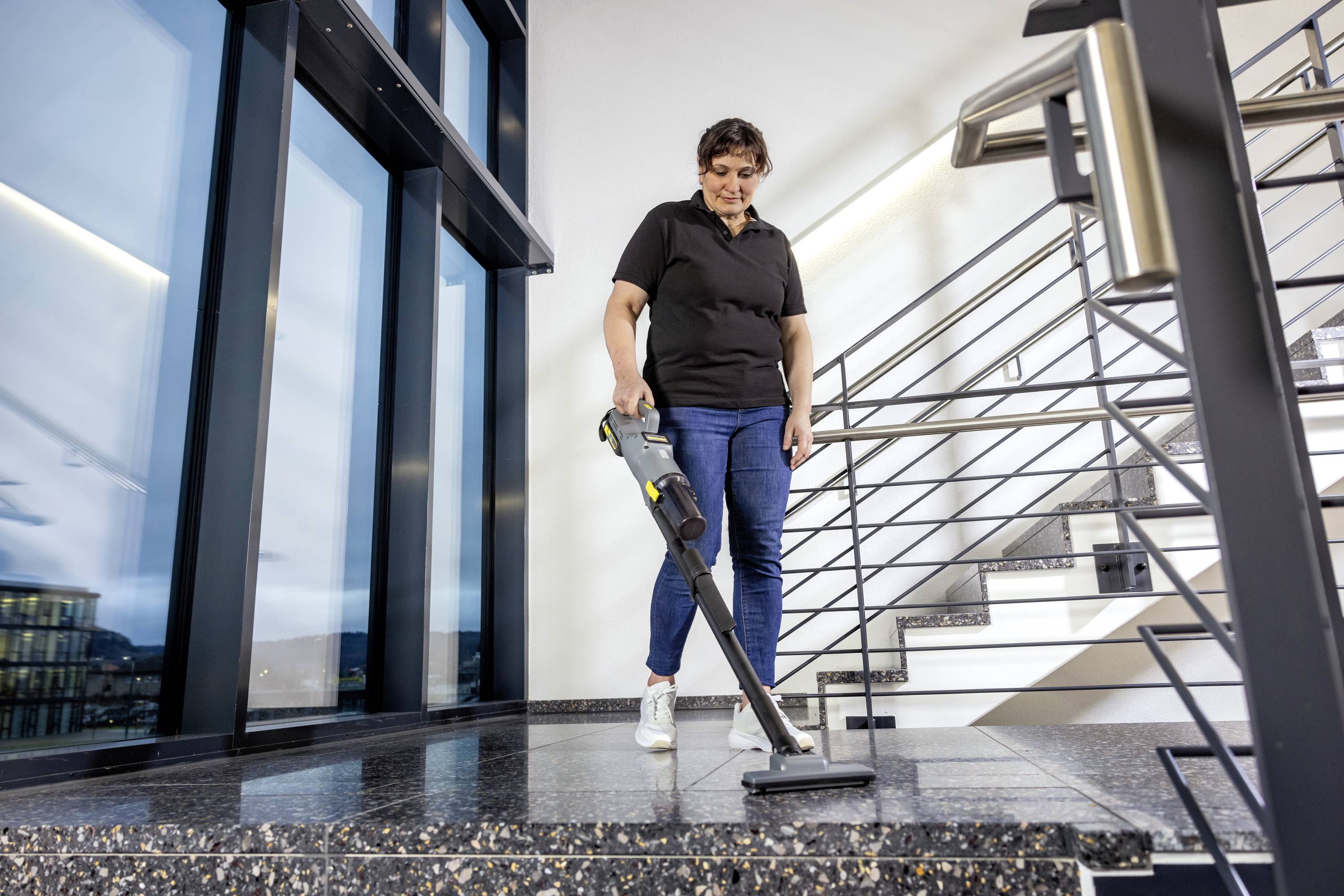 Eine Person saugt mit einem kabellosen Staubsauger einen Treppenabsatz in einem modernen Gebäude. Der Bereich verfügt über große Fenster und Metallgeländer.