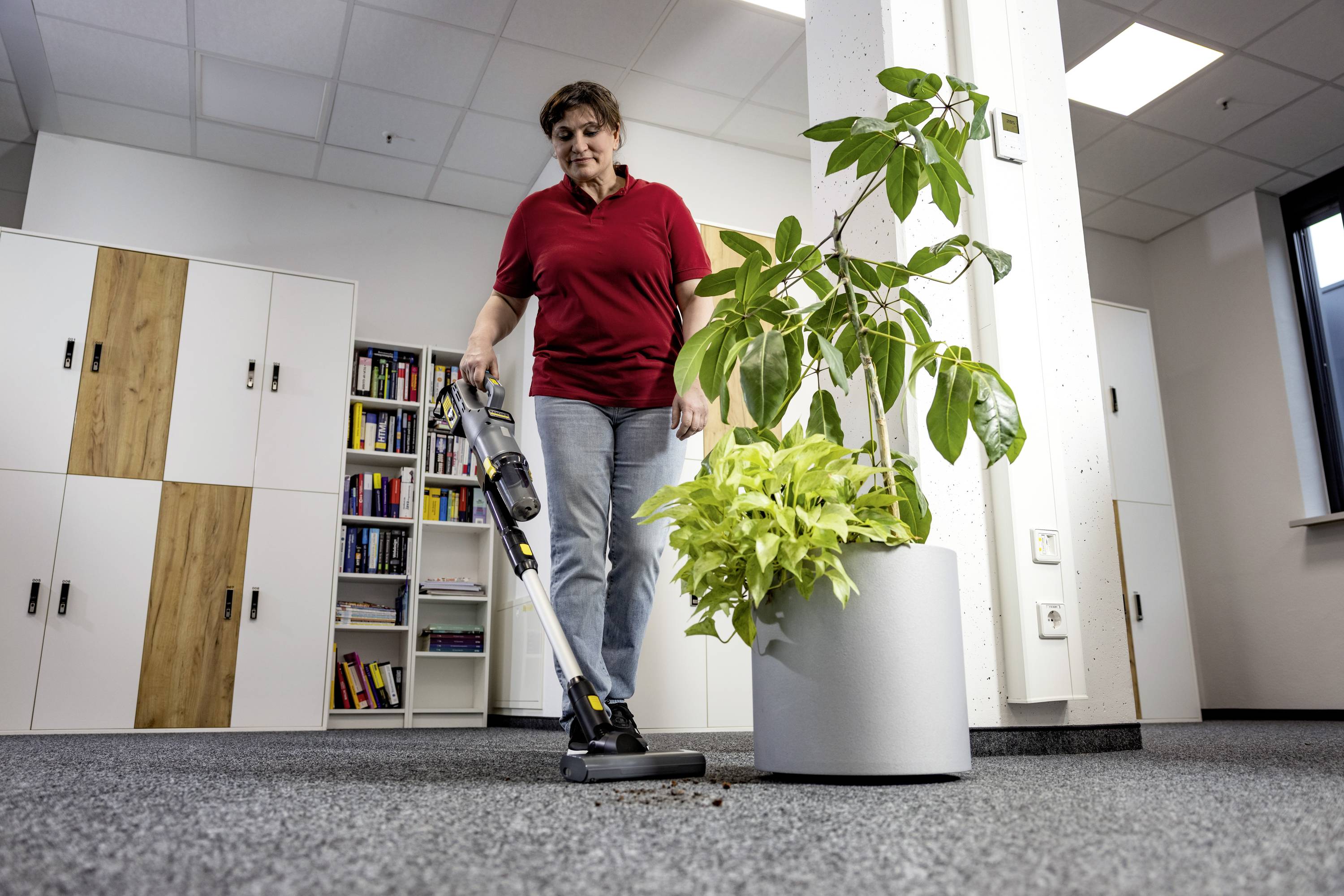 Eine Person saugt einen Teppichboden in einem Büroraum mit einem kabellosen Staubsauger, während im Hintergrund Blumentöpfe und Bücherregale zu sehen sind.