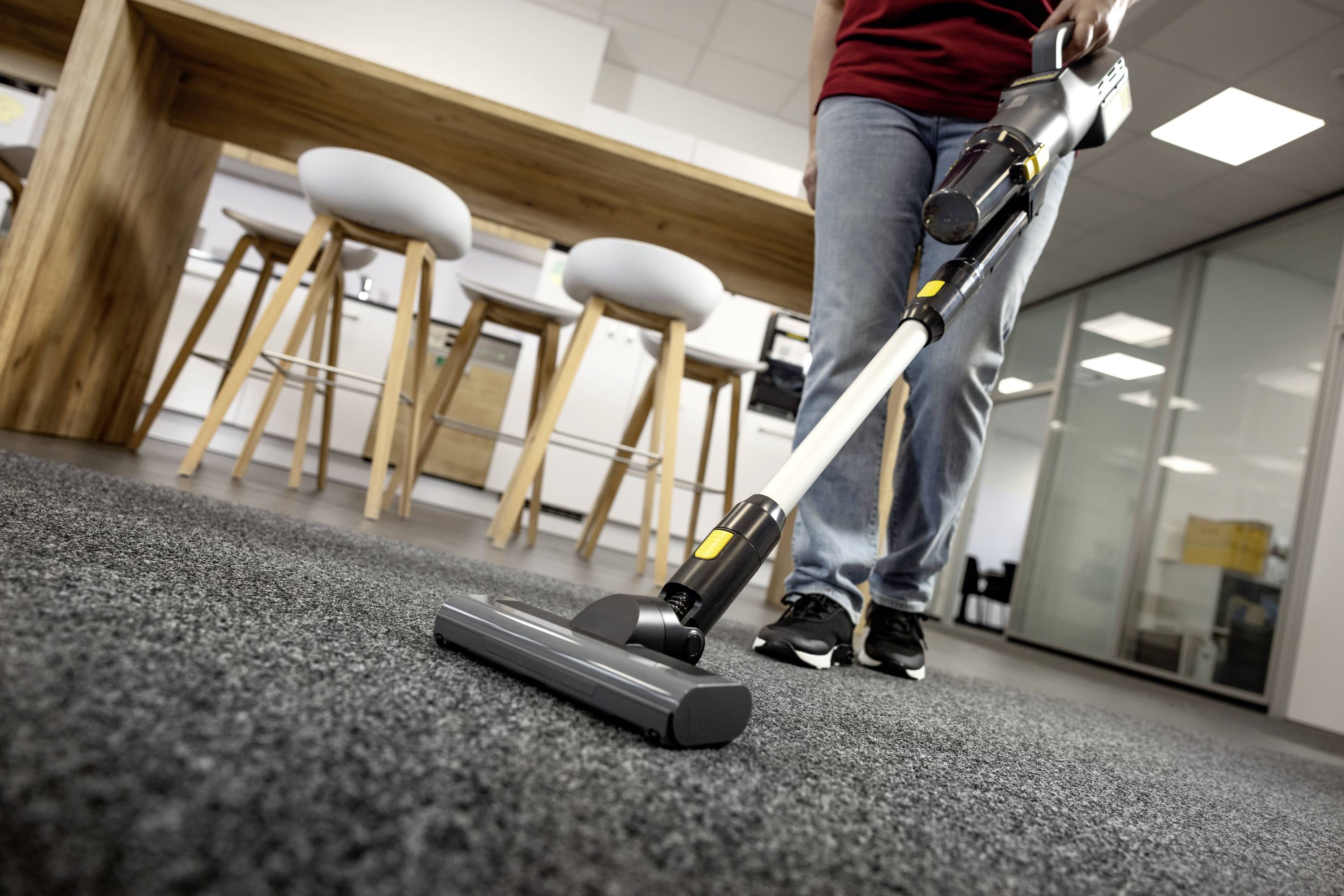 Eine Person, die einen Teppichboden in einem modernen Büroraum mit Hockern und einem Holztisch im Hintergrund staubsaugt.