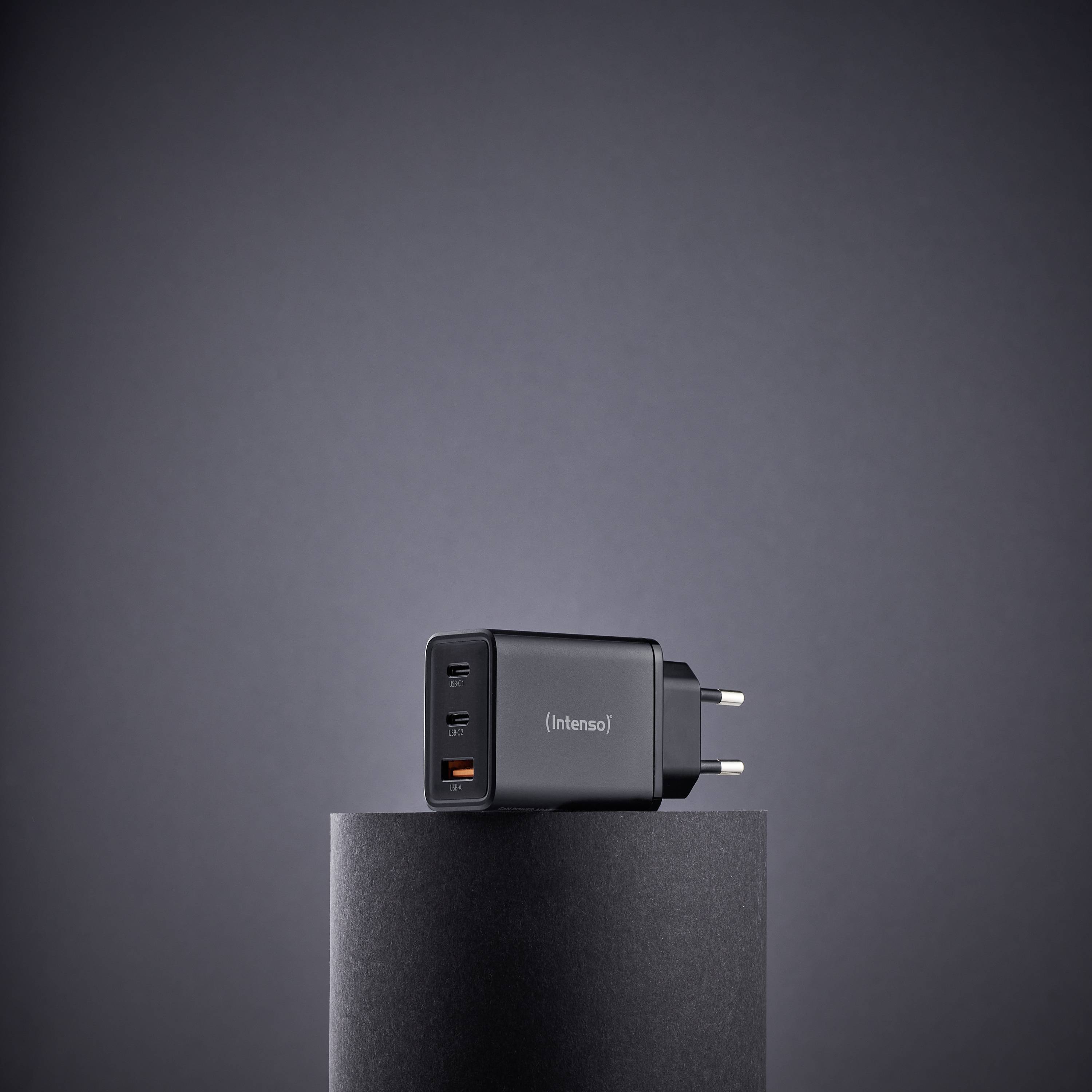 Ein schwarzer USB-Wandladegerät mit mehreren Anschlüssen steht auf einem zylindrischen Sockel vor einem einfarbigen grauen Hintergrund.