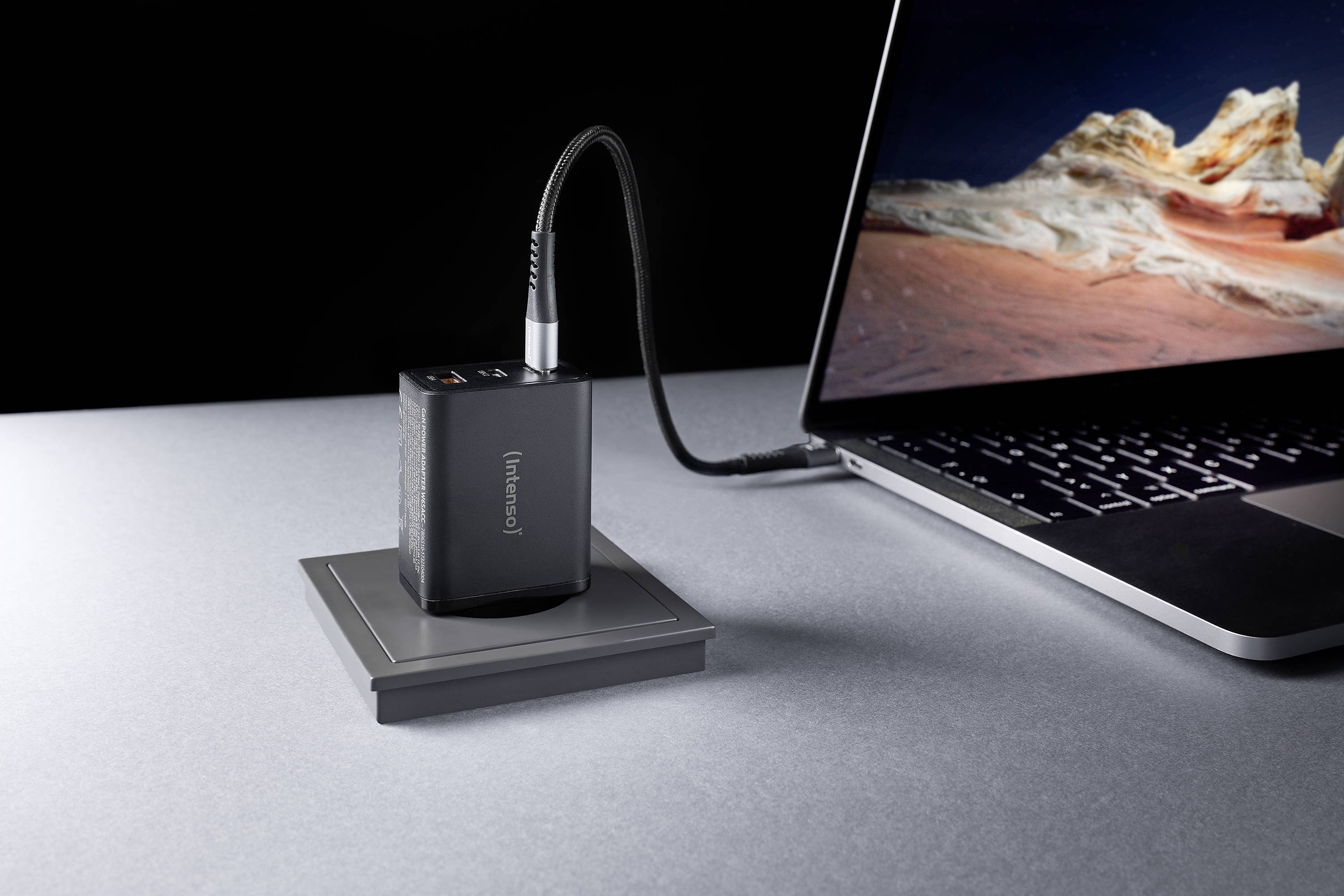 Eine tragbare Powerbank lädt einen Laptop auf einem grauen Schreibtisch. Der Laptop-Bildschirm zeigt ein Landschaftsbild.