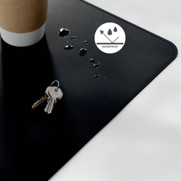 Eine Kaffeetasse steht auf einer schwarzen, wasserdichten Matte, auf der sich Wassertropfen bilden. Schlüssel liegen in der Nähe und deuten auf eine Widerstandsfähigkeit gegen Verschüttungen hin.