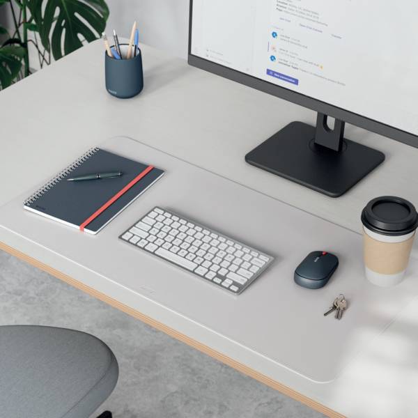 Ein minimalistischer Arbeitsplatz mit einem Computermonitor, Tastatur, Notizbuch, Stiften, einer Kaffeetasse und einer Maus auf einem hellen Holzschreibtisch.