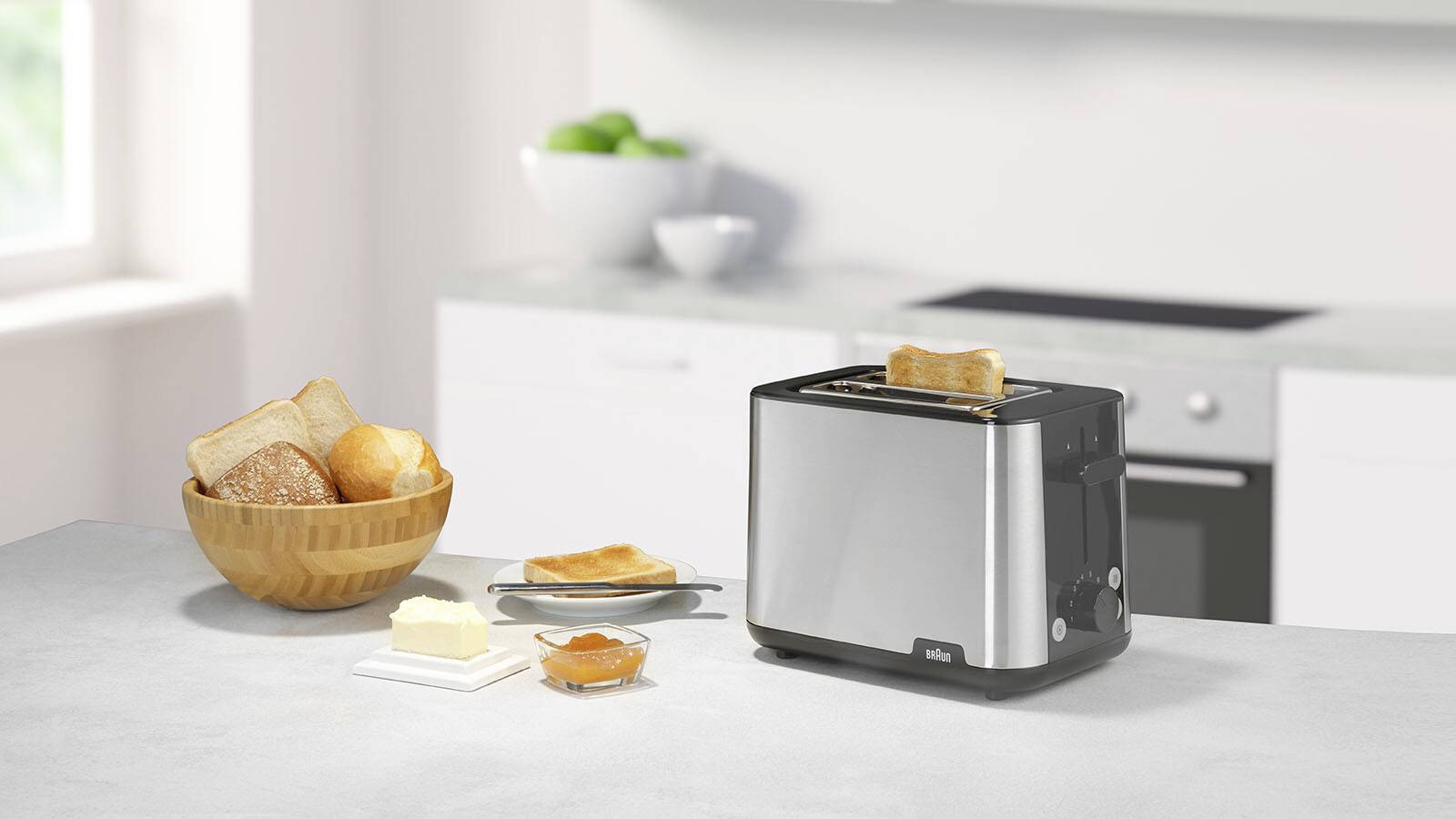 Ein Toaster aus Edelstahl auf einer Küchenarbeitsplatte, der ein Stück Brot röstet. Daneben eine Schüssel mit Brot, Butter und Marmelade auf einem Teller.