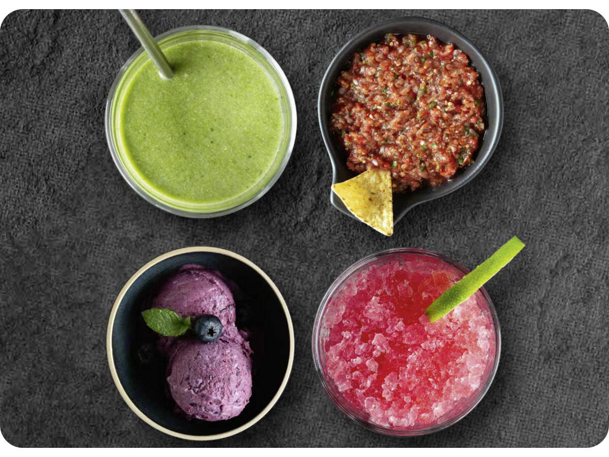 Vier Schalen auf einer dunklen Oberfläche, die einen grünen Smoothie, rote Salsa mit einem Tortillachip, lila Eis mit Beeren und einen pinken Slush mit einer Limette enthalten.
