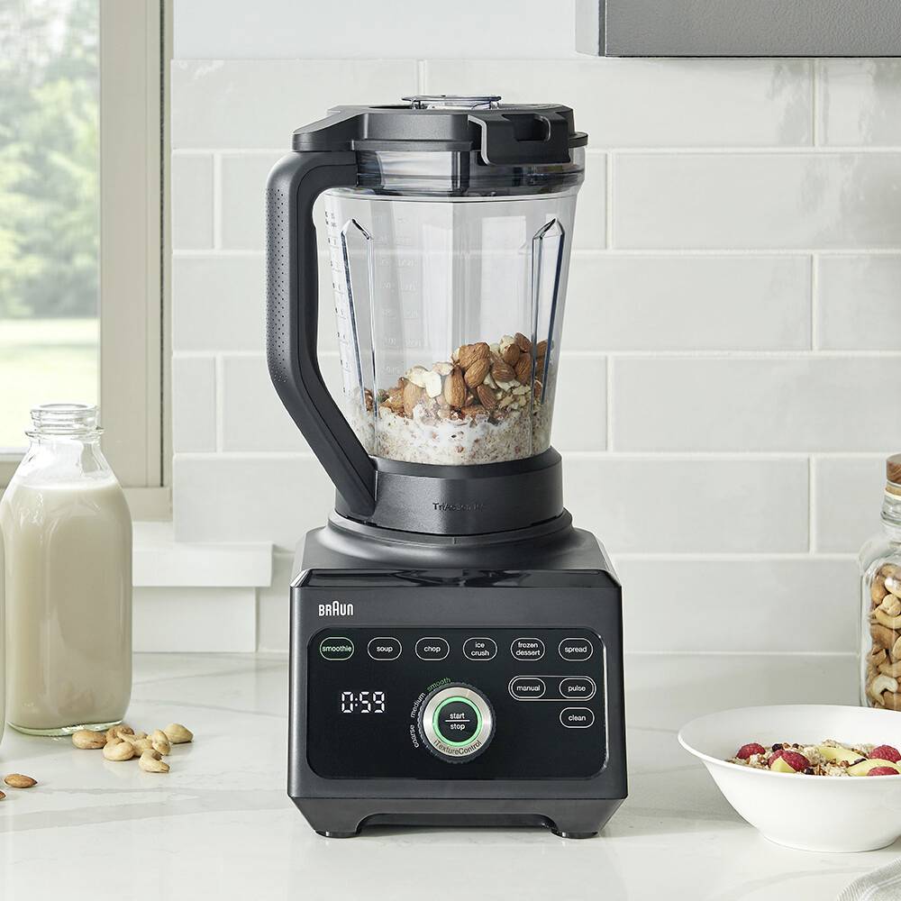 Ein Mixer auf einer Küchenarbeitsplatte, gefüllt mit Mandeln und Haferflocken. In der Nähe stehen eine Milchflasche und ein Glas, was auf die Zubereitung eines Smoothies hindeutet.