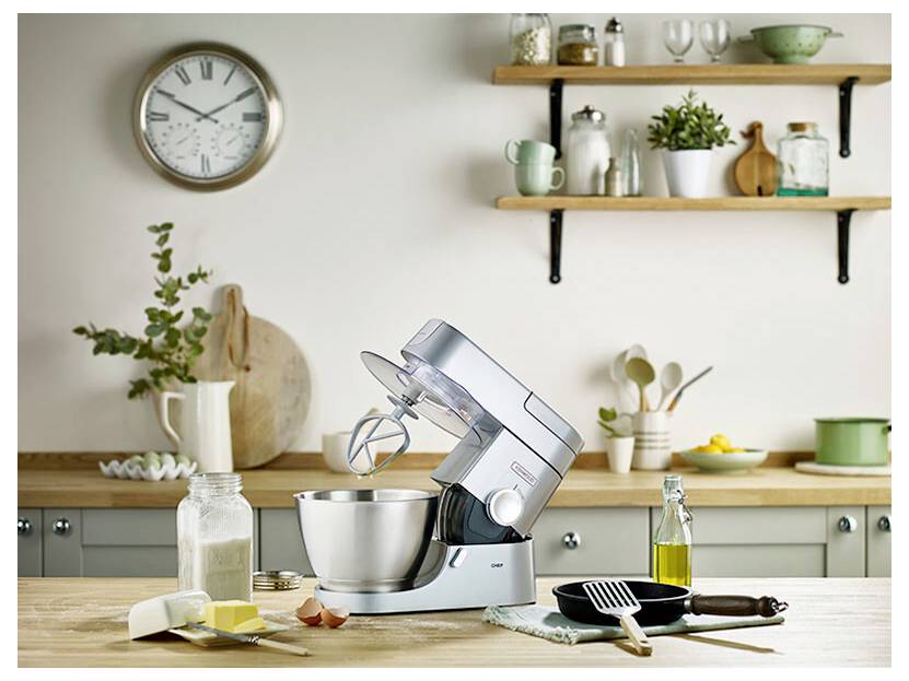 Eine Küchenarbeitsplatte mit einem Standmixer, Backzutaten und Küchenutensilien im Vordergrund; Regale mit Geschirr und einer Uhr im Hintergrund.