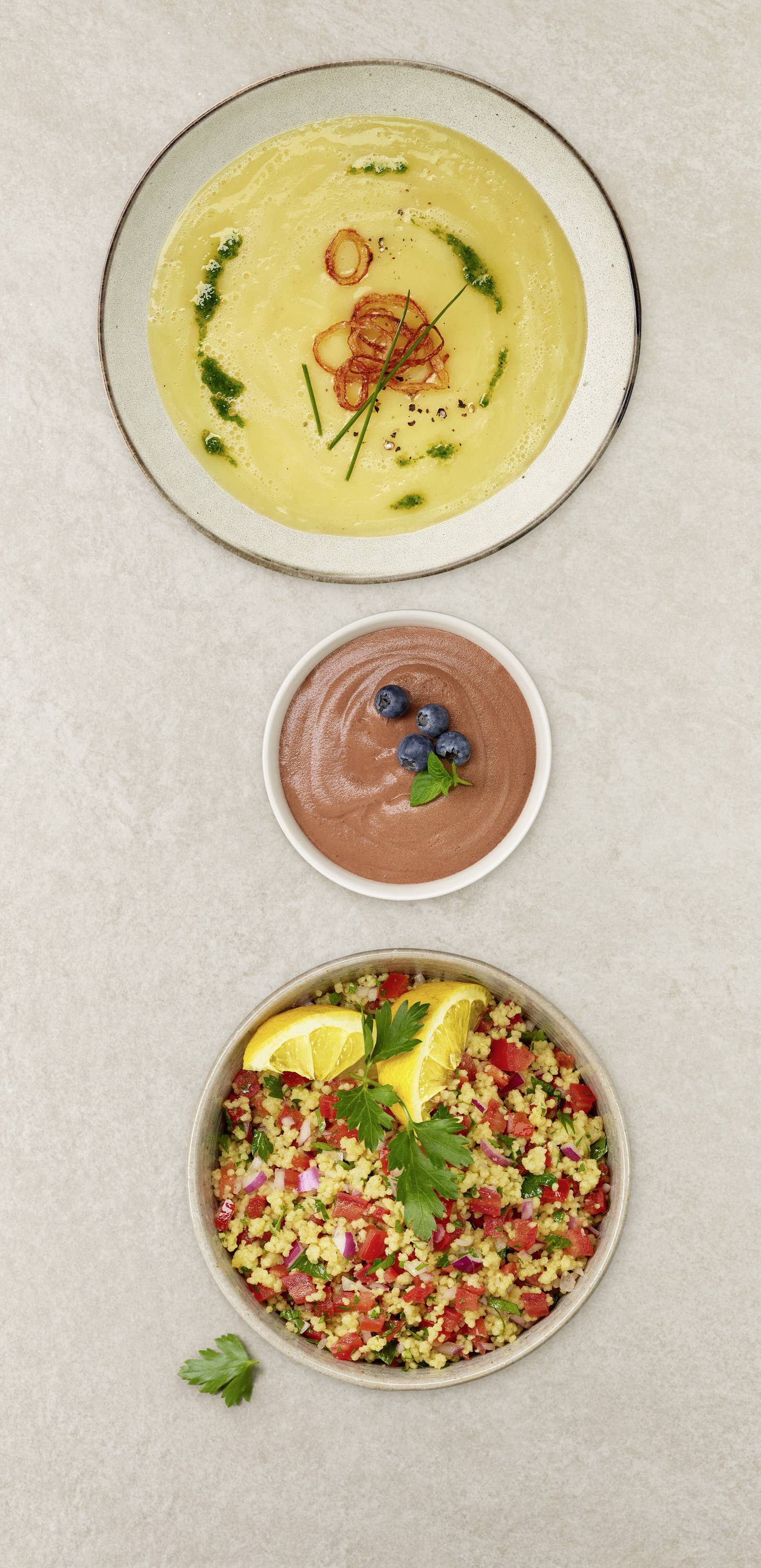 Eine Tafel zeigt drei Gerichte: eine Schüssel gelbe Suppe garniert mit Kräutern, eine kleine Schüssel Schokoladenmousse mit Blaubeeren und einen Teller bunten Couscous-Salat mit Zitronenscheiben.