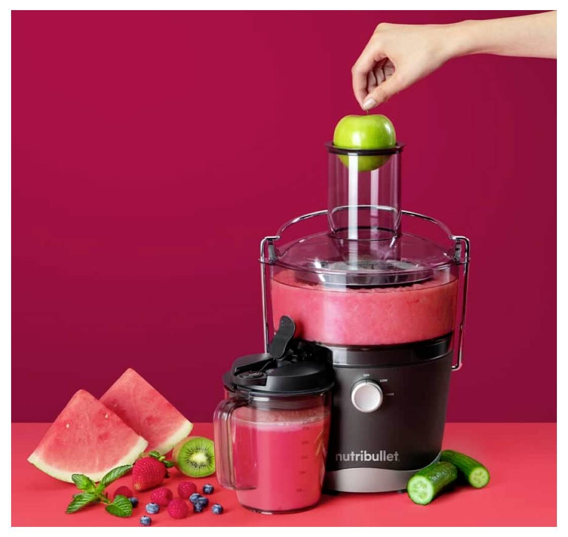 Eine Hand legt einen grünen Apfel in einen Nutribullet-Entsafter mit rotem Saft, umgeben von Wassermelone, Beeren, Gurke und Kiwi.