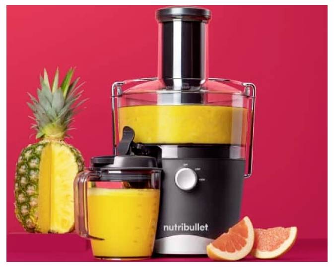 Eine schwarze und silberne Nutribullet-Entsaftungsmaschine, gefüllt mit Orangensaft, steht auf einem roten Hintergrund. Daneben liegen eine aufgeschnittene Ananas und zwei Grapefruit-Spalten.