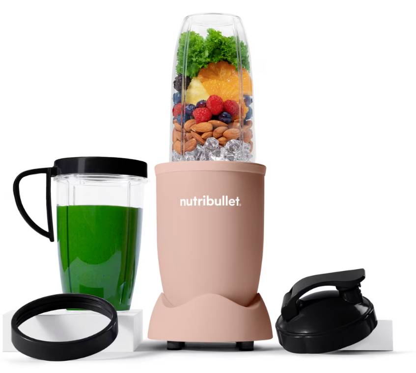 Ein pinker NutriBullet-Mixer mit Früchten, Mandeln und Grünkohl in der Tasse. In der Nähe befinden sich ein grünes Getränk in einer To-Go-Tasse, ein Deckel und ein Dichtungsring.