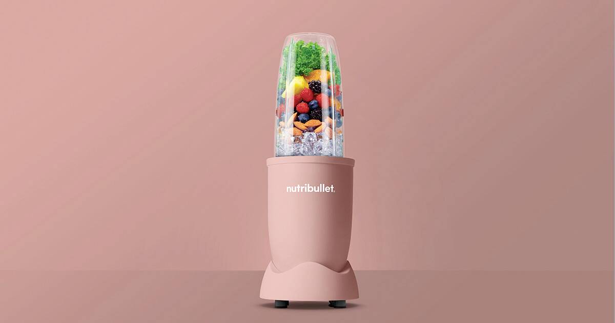 Ein pinkfarbener NutriBullet-Mixer, gefüllt mit verschiedenen Obst- und Gemüsesorten, darunter Beeren und Blattgemüse, vor einem passenden pinkfarbenen Hintergrund.