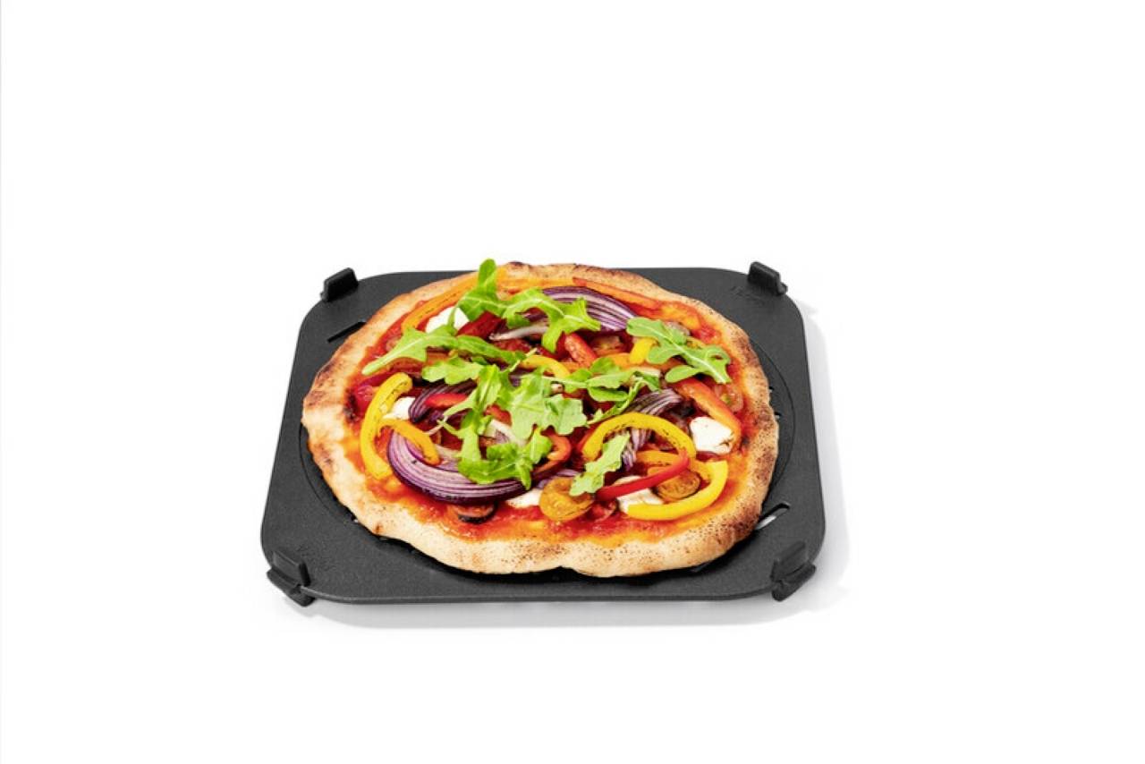 Eine Pizza belegt mit roten Zwiebeln, gelben Paprikaschoten und Rucola auf einem schwarzen Backblech vor einem schlichten weißen Hintergrund.