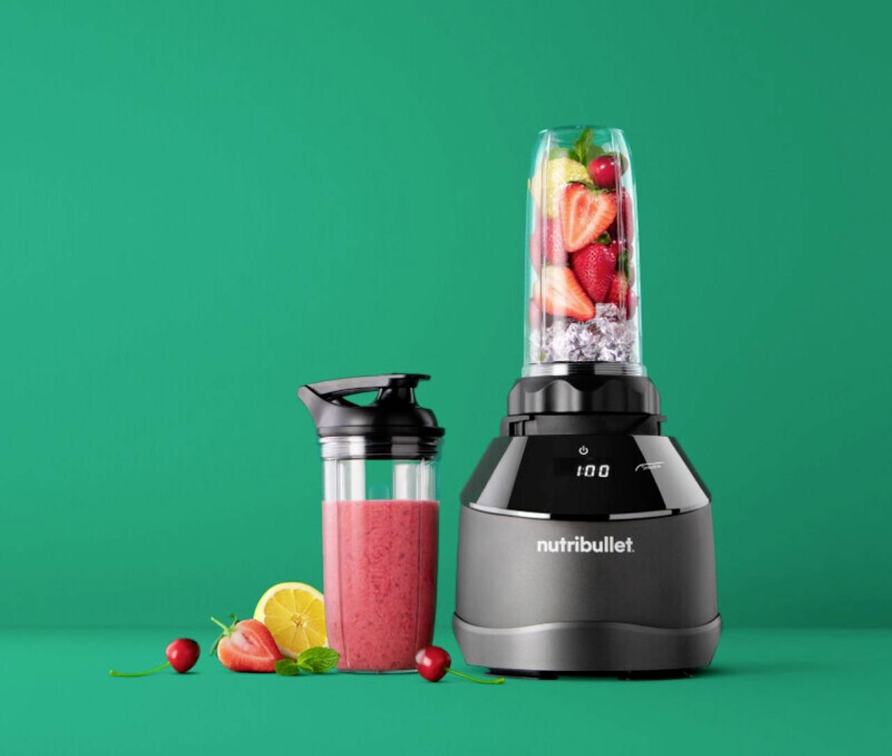Ein Mixer voller Früchte neben einer Smoothie in einer Flasche, vor einem grünen Hintergrund. Die Marke 'Nutribullet' ist sichtbar.