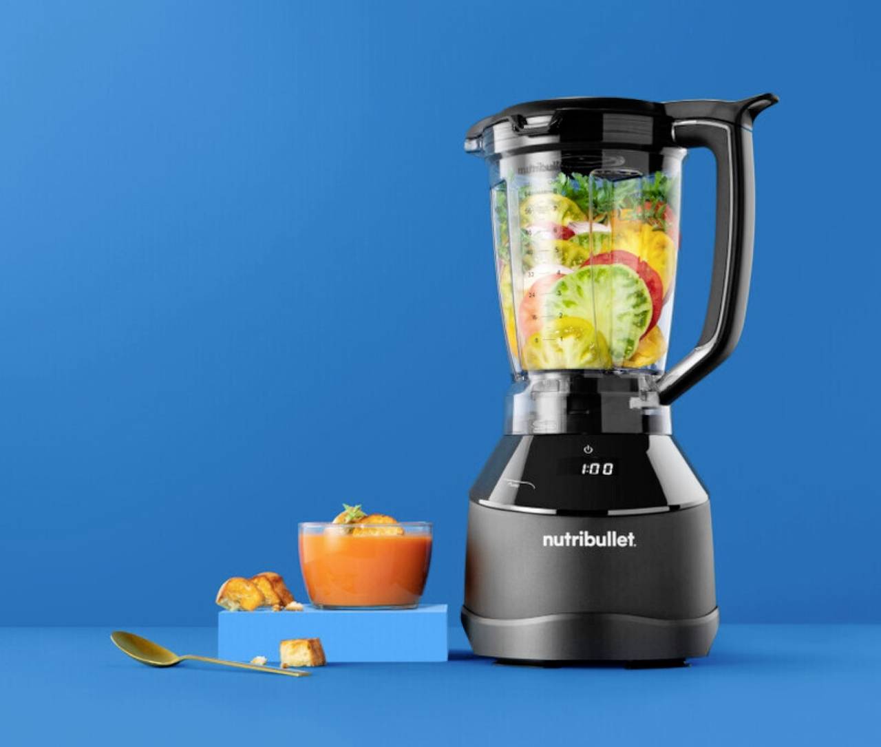 Ein Mixer, gefüllt mit verschiedenen Obst- und Gemüsesorten, steht neben einer Schüssel Suppe und einem Löffel, vor einem hellblauen Hintergrund.