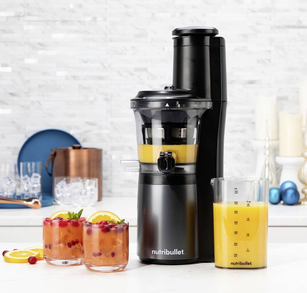 Ein schwarzer Nutribullet-Entsafter auf einer Küchenarbeitsplatte mit zwei Gläsern bunter Fruchtsäfte und einem Krug mit Orangensafe daneben.