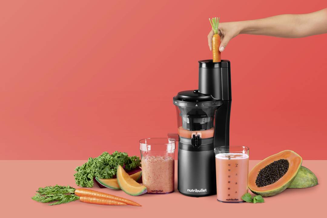 Eine Hand legt eine Karotte in einen Nutribullet-Entsafter, umgeben von Grünkohl, Karotten, Papaya und Gläsern Orangensafts vor einem roten Hintergrund.