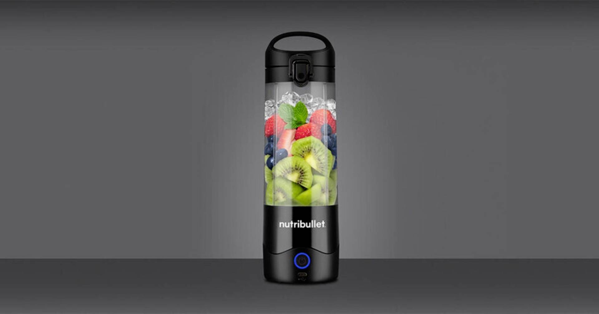 Eine tragbare Blenderflasche gefüllt mit verschiedenen Früchten, darunter Kiwi, Erdbeeren und Blaubeeren, vor einem grauen Hintergrund.