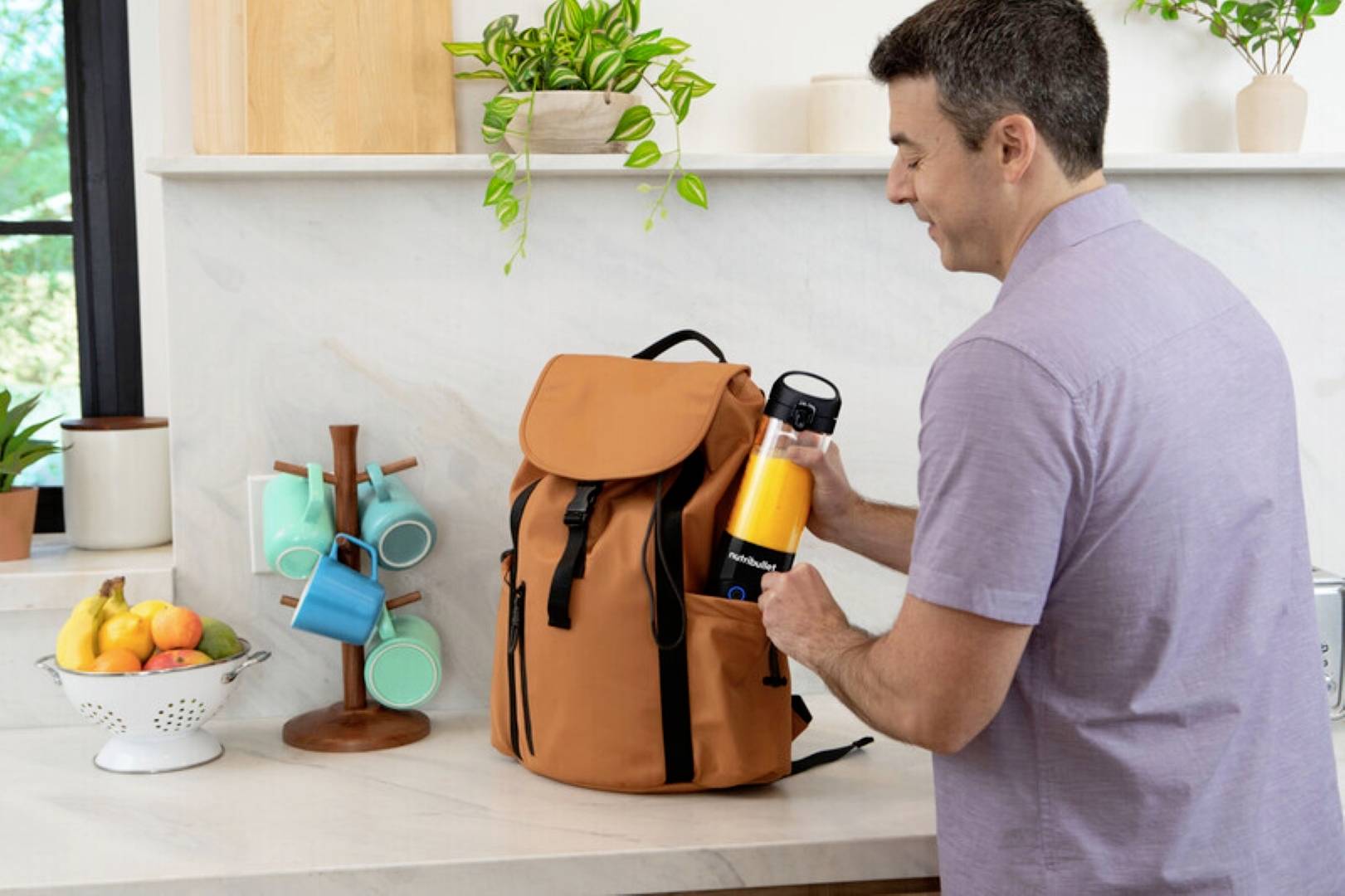 Ein Mann legt eine Wasserflasche in einen braunen Rucksack auf eine Küchenarbeitsplatte. In der Nähe sind gestapelte Tassen und eine Schüssel mit Obst.