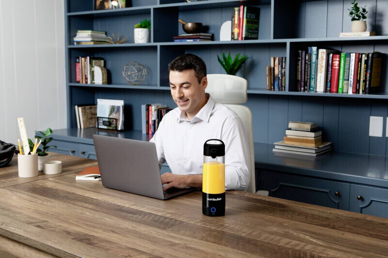 Mann arbeitet an einem Laptop an einem Holzschreibtisch, mit einem tragbaren gelben Luftreinigungsgerät in der Nähe. Der Hintergrund zeigt ein blaues Bücherregal mit Büchern und Dekorationen.