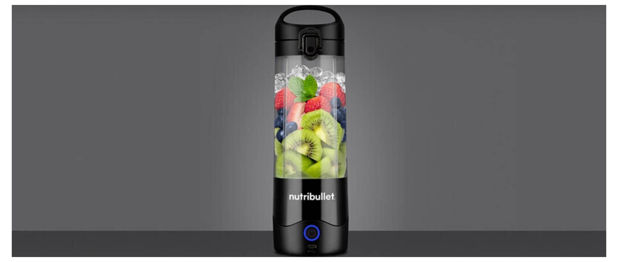 Ein schwarzer Nutribullet-Mixer-Behälter, gefüllt mit Kiwi, Erdbeeren, Blaubeeren und Blattgrün vor einem grauen Hintergrund.