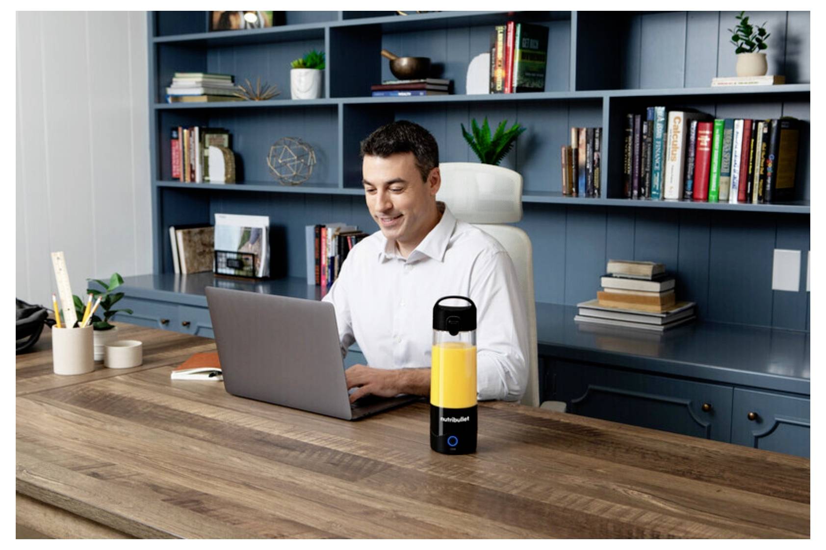 Eine Person sitzt an einem Schreibtisch und arbeitet an einem Laptop in einem modernen Büro mit einem Bücherregal im Hintergrund. Eine gelbe Wasserflasche steht auf dem Schreibtisch.