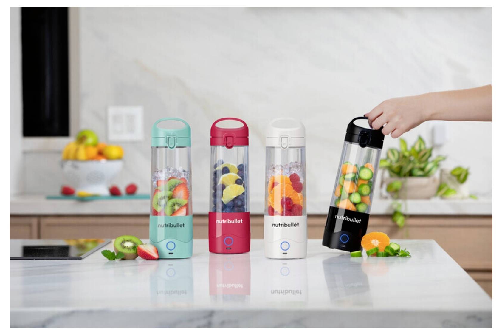 Vier buonte Nutribullet-Mixer mit Früchten in jedem stehen auf einer Küchenarbeitsplatte. Eine Hand greift nach dem schwarzen Mixer. Im Hintergrund befindet sich eine Obstschale und eine Pflanze.