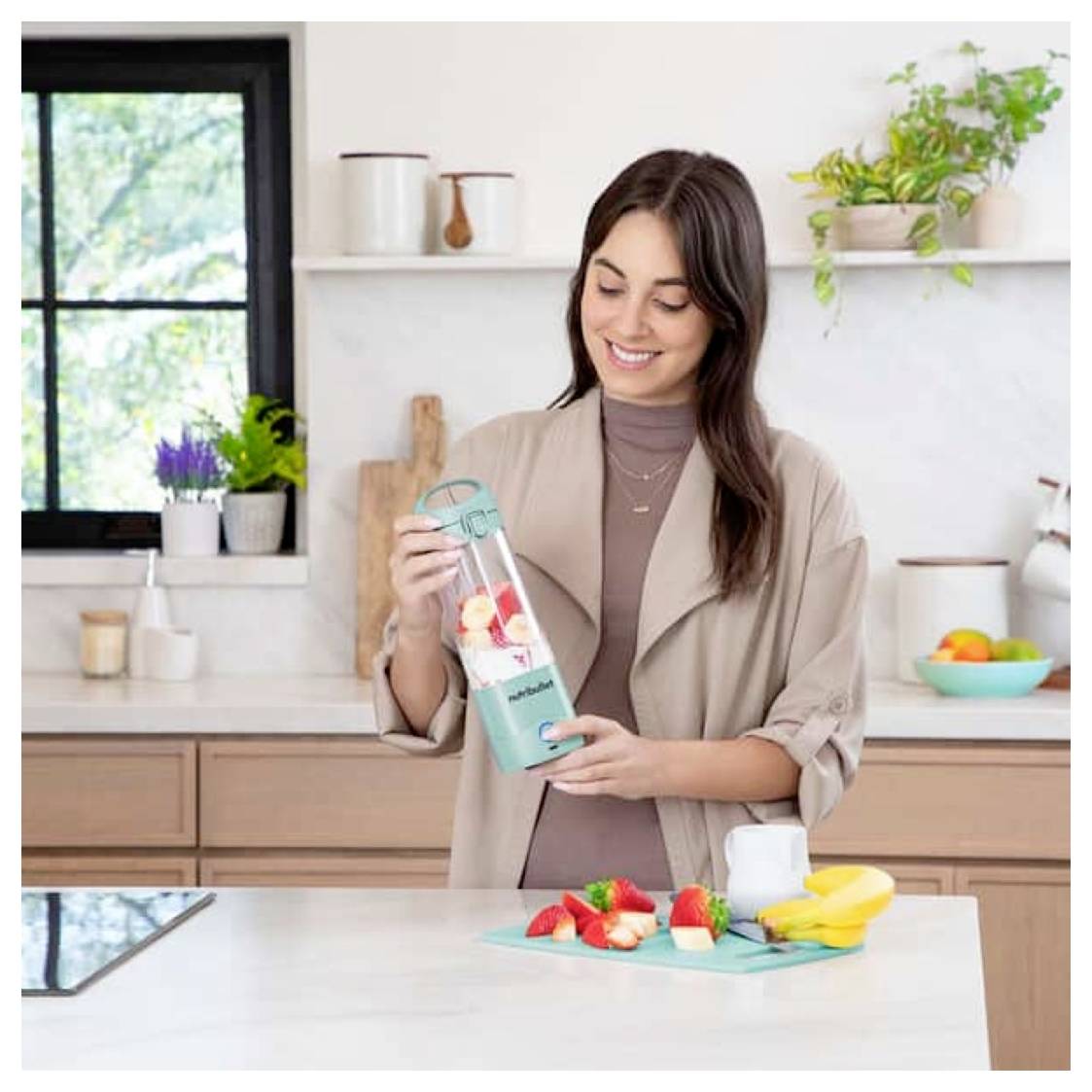 Eine Person in einer Küche lächelt, während sie einen Smoothie mit einem tragbaren Mixer zubereitet, der mit Obst gefüllt ist. Ein Brett mit Obst steht auf der Arbeitsplatte.