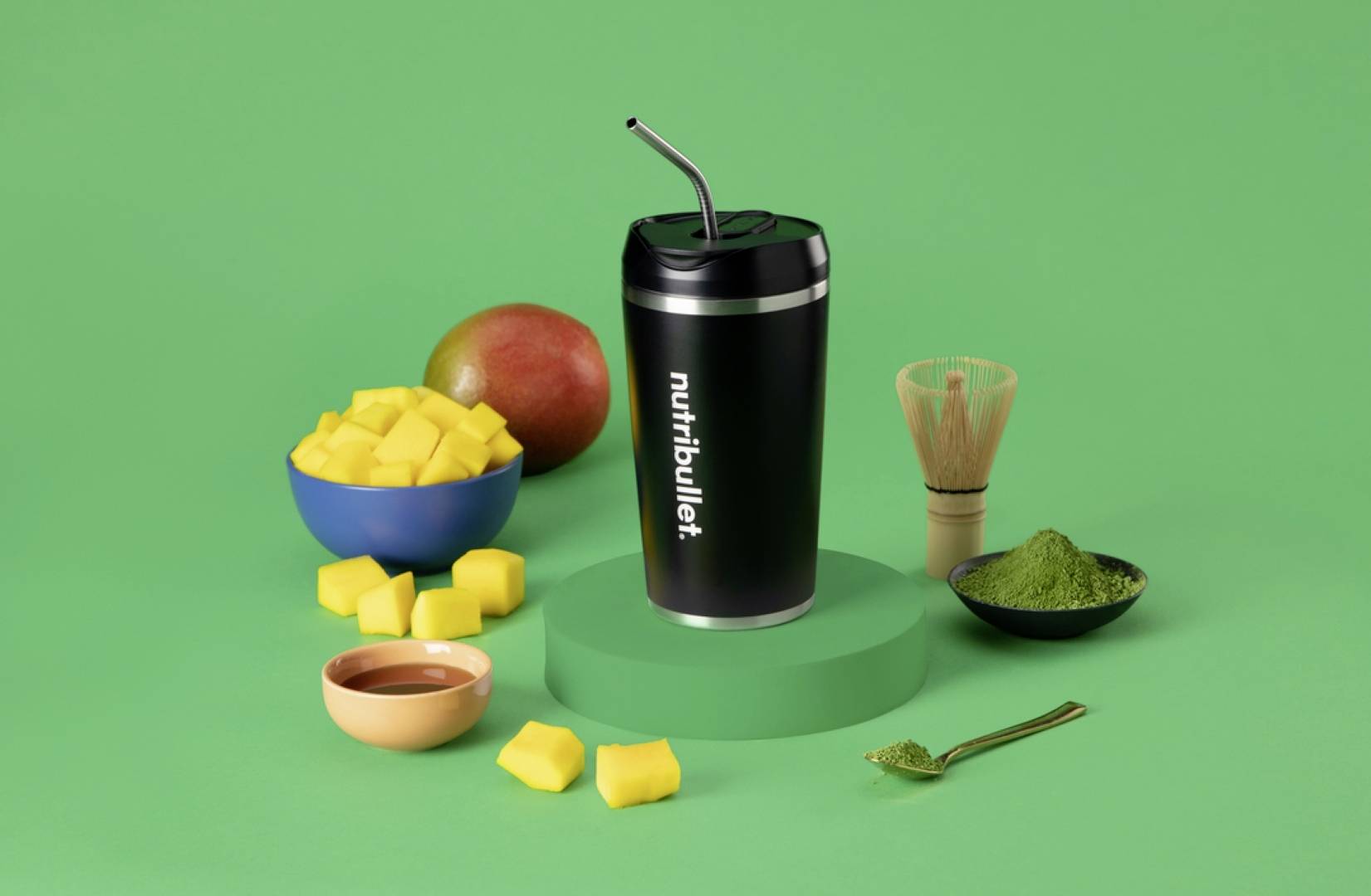 Ein schwarzer Nutribullet-Becher mit einem Strohhalm auf einer grünen Plattform, umgeben von gewürfelter Mango, einer ganzen Mango, Matcha-Pulver und einem Schneebesen.