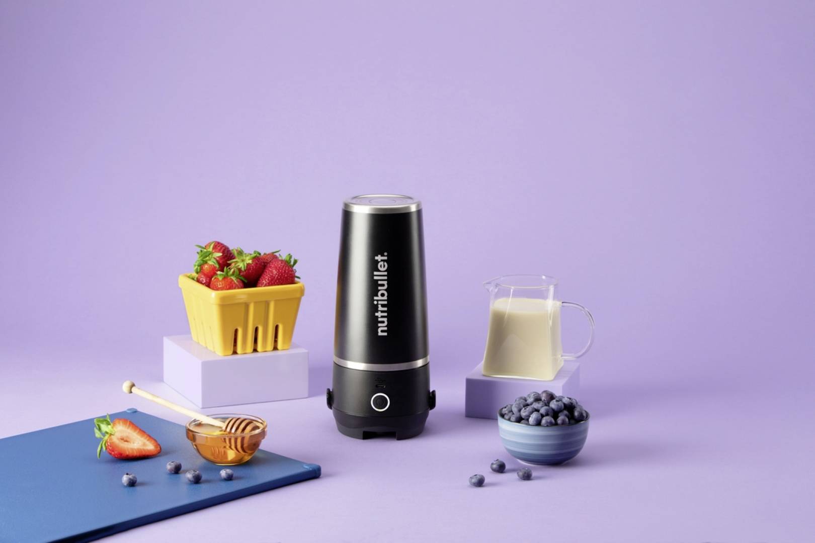 Ein Mixer mit 'Nutribullet'-Branding ist von Erdbeeren, Milch und Blaubeeren vor einem lila Hintergrund umgeben, was die Zubereitung eines Smoothies andeutet.