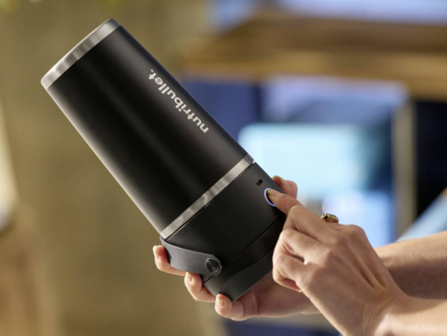 Eine Person, die einen schlanken schwarzen tragbaren Mixer hält und einen Knopf drückt. Das Gerät ist mit 'nutribullet' beschriftet. Der Hintergrund ist weich unscharf.
