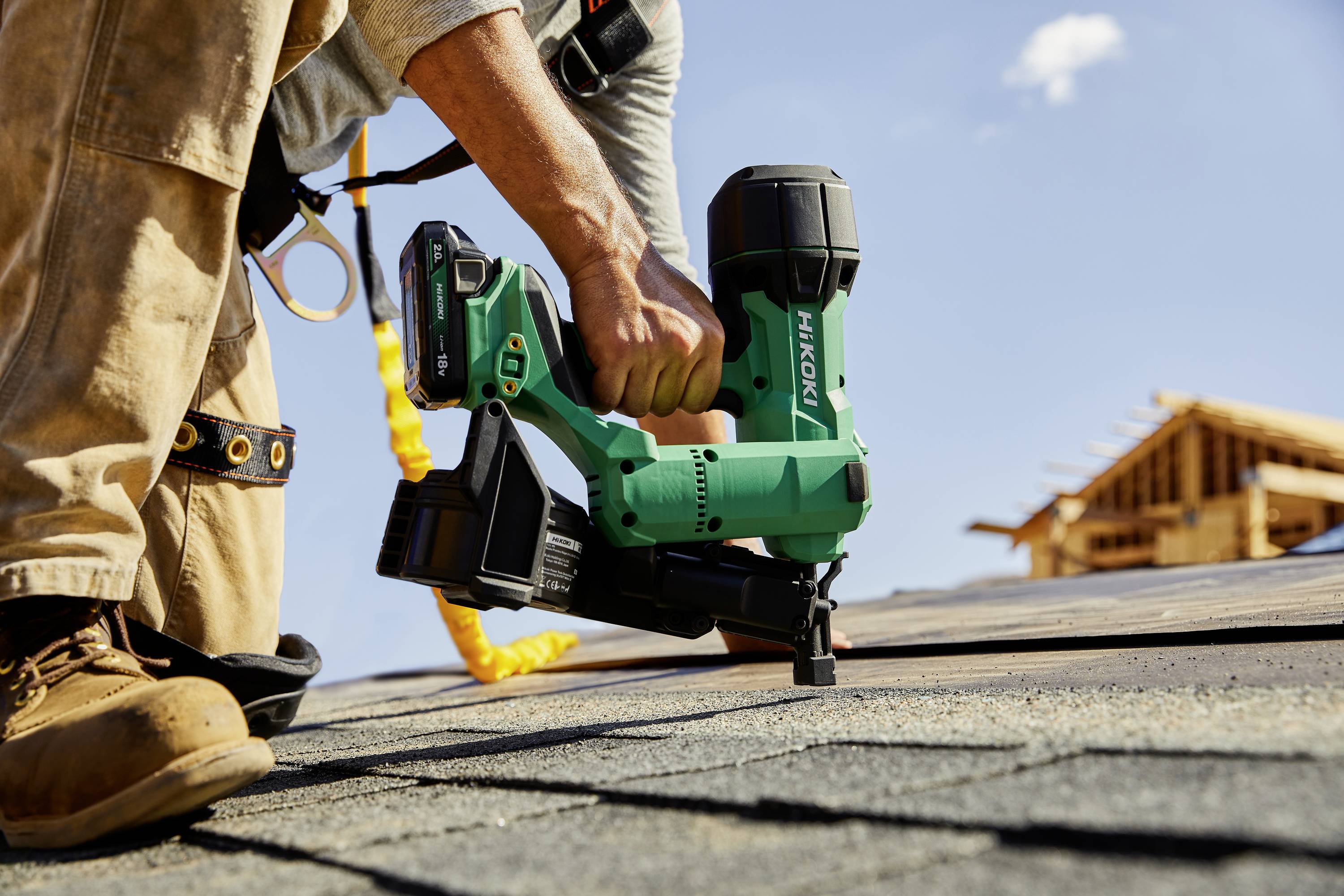 Ein Bauarbeiter in braunen Stiefeln und Sicherheitsausrüstung benutzt ein grünes Elektrowerkzeug auf einem Holzdach unter einem klaren blauen Himmel.