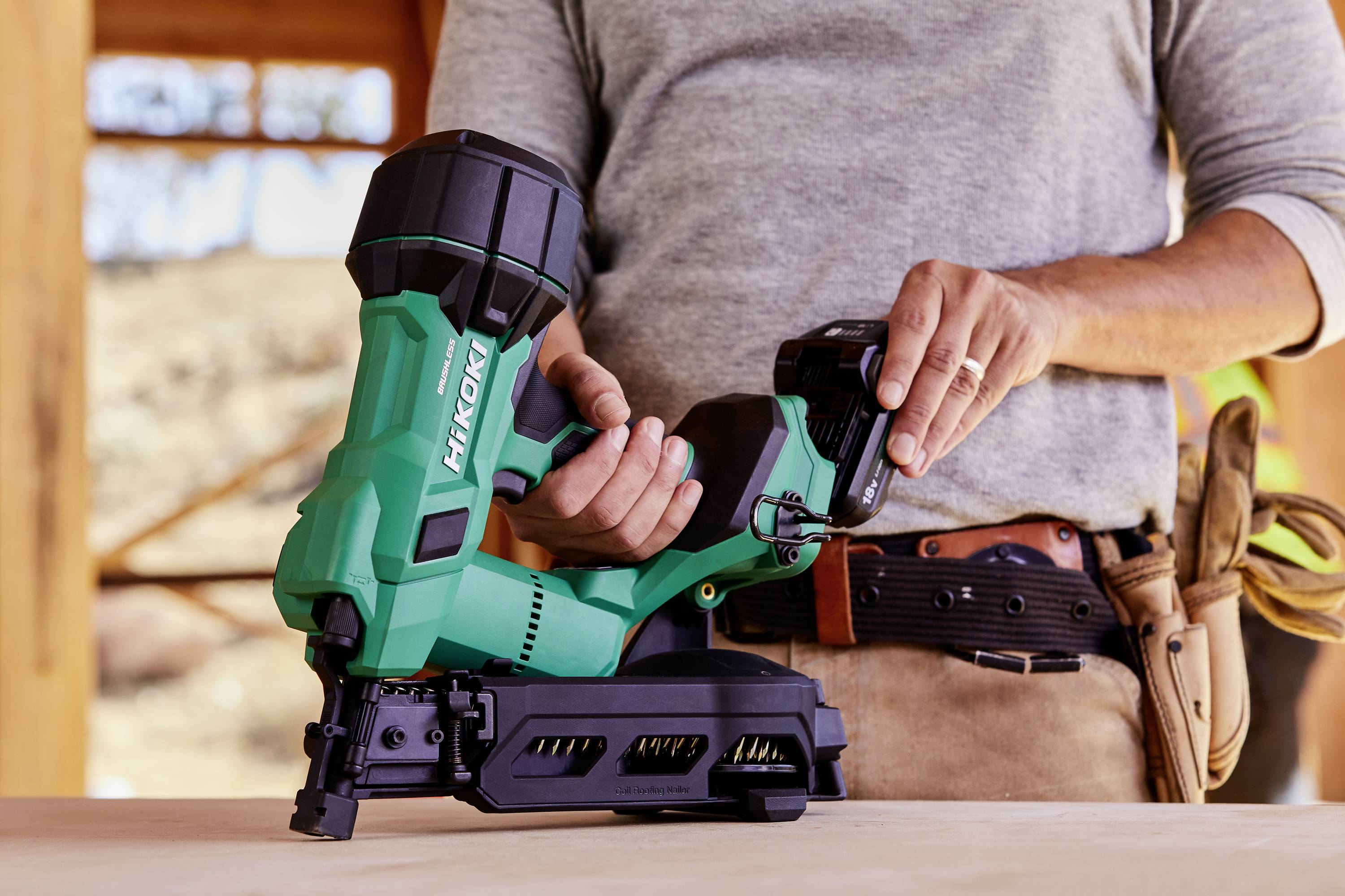 Eine Person, die ein grünes Elektrowerkzeug, möglicherweise einen Nagler, in einer Werkstattumgebung mit Holzbalken hält; trägt einen Werkzeuggürtel und lässige Kleidung.